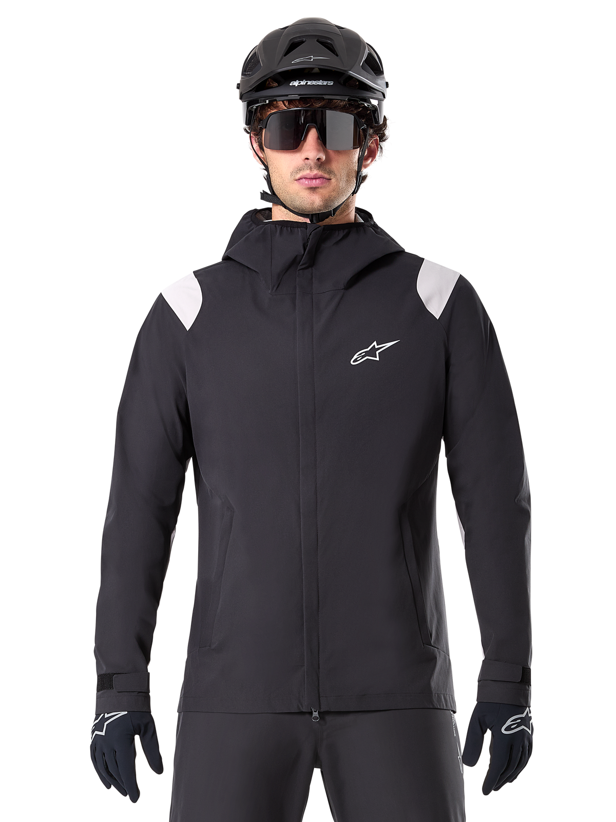 Alpinestars A-Dura regnjacka, MTB-regnjacka, svart med vita axelpaneler, bärs med svart MTB-hjälm, solglasögon och handskar, vattentät design med huva och dragkedja fram, sportig friluftsutrustning för cykling