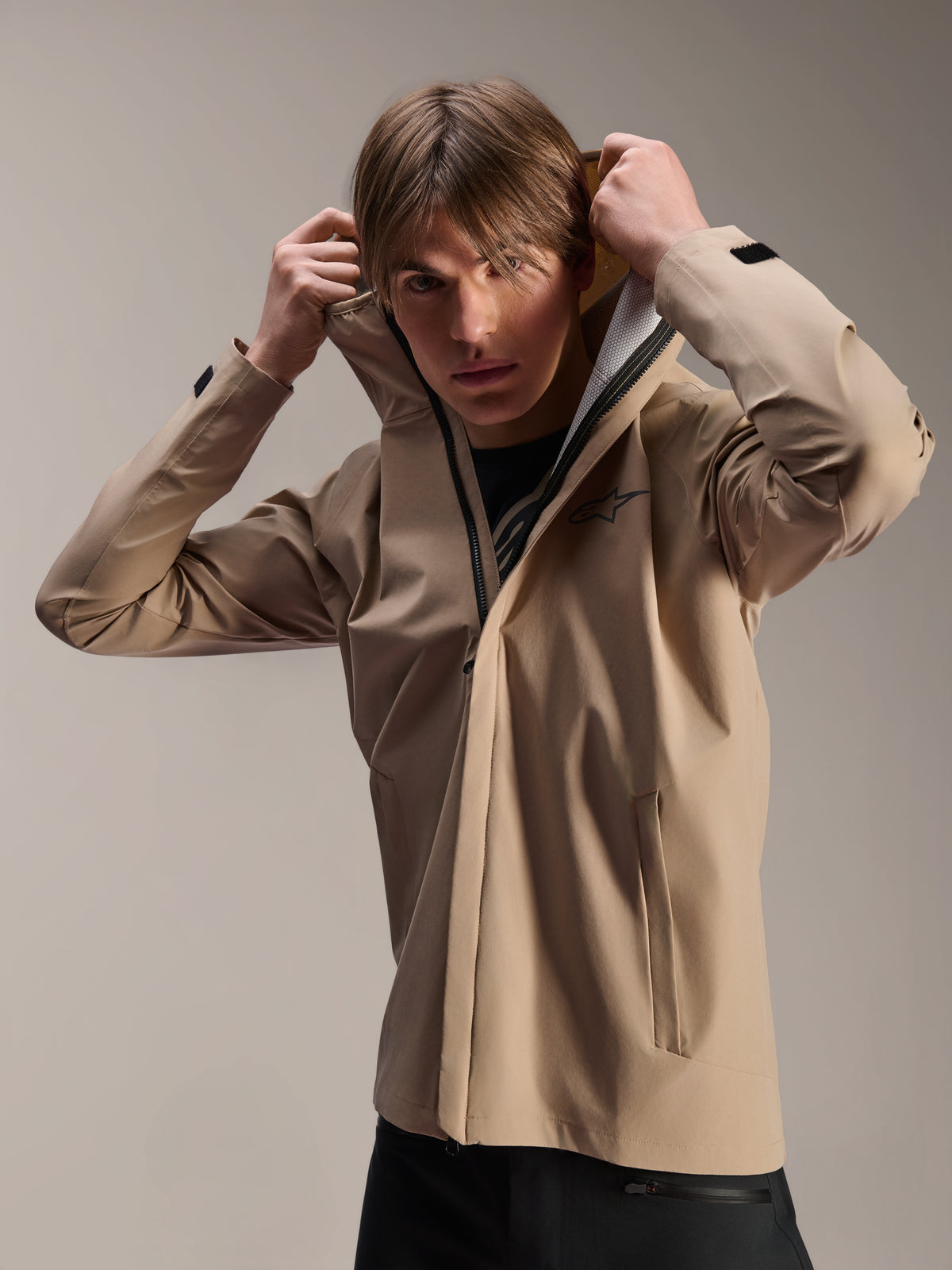 Alpinestars A-Dura Rain Jacket, MTB-cykeljacka, ljusbrun, modell som bär jackan och drar upp den elastiska huvan, med en svart logotyp på bröstet, fickor med dragkedja och tekniskt vattentätt 2,5-lagersmaterial