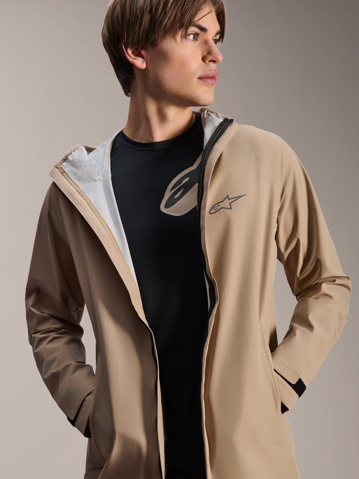 Manlig modell bär Alpinestars A-Dura regnjacka, ljusbrun, öppen över en svart t-shirt, visar vitt meshfoder, mångsidig väderbeständig cykeljacka