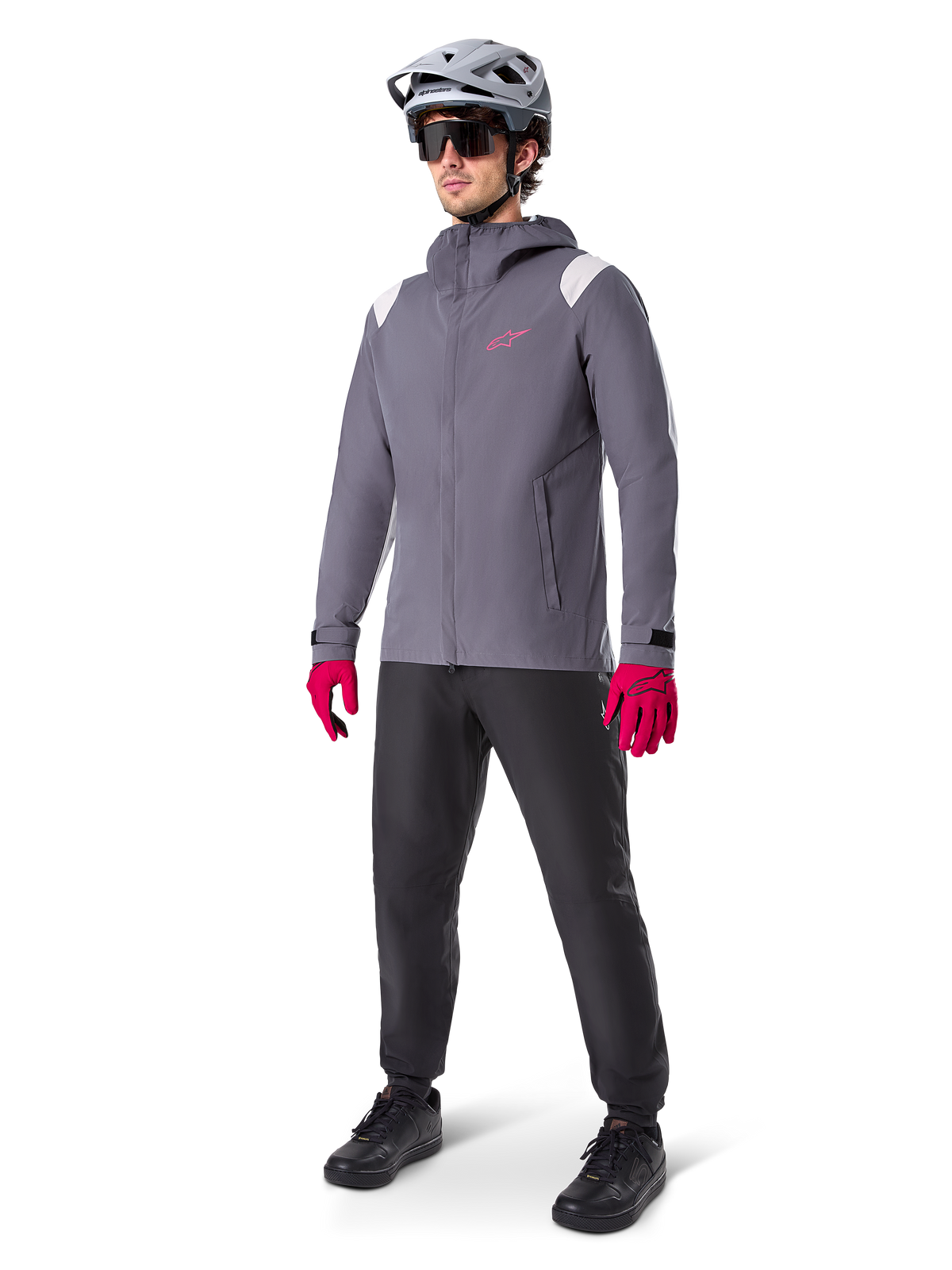 Manlig modell som bär Alpinestars A-Dura Regnjacka i Dark Titanium, grå jacka med huva med rosa logotyp på bröstet och ljusgrå axelpaneler, matchad med en grå MTB-hjälm, mörka solglasögon, starkt rosa handskar och svarta byxor för terrängcykling.