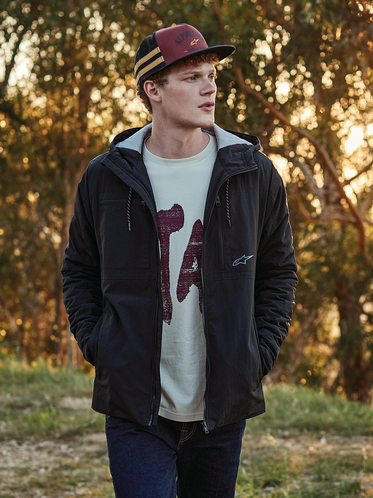 Alpinestars Backline Trucker Hat i Brick, svart och senapsgult, bärs av en man kombinerat med en svart huvjacka och grafisk t-shirt, avslappnad motorsportinspirerad streetwear i en utomhusmiljö under den gyllene timmen.