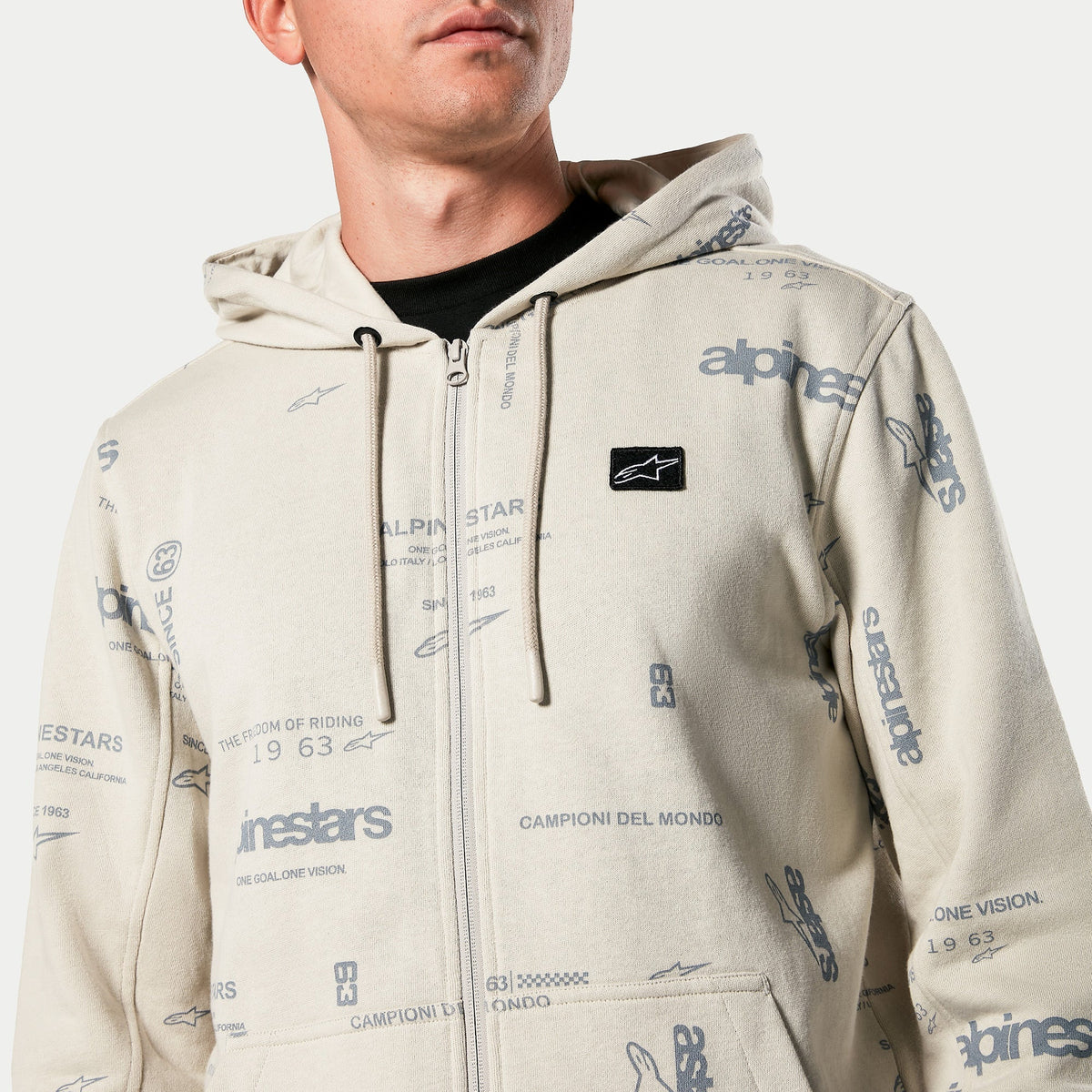 Alpinestars Plenitude Hoodie, fleecehoodie med dragkedja fram i Off White, heltäckande grafiskt tryck med texten 'alpinestars' och '1963', svart broderat logomärke på bröstet, huva med dragsko, casual motorsportinspirerad streetwear