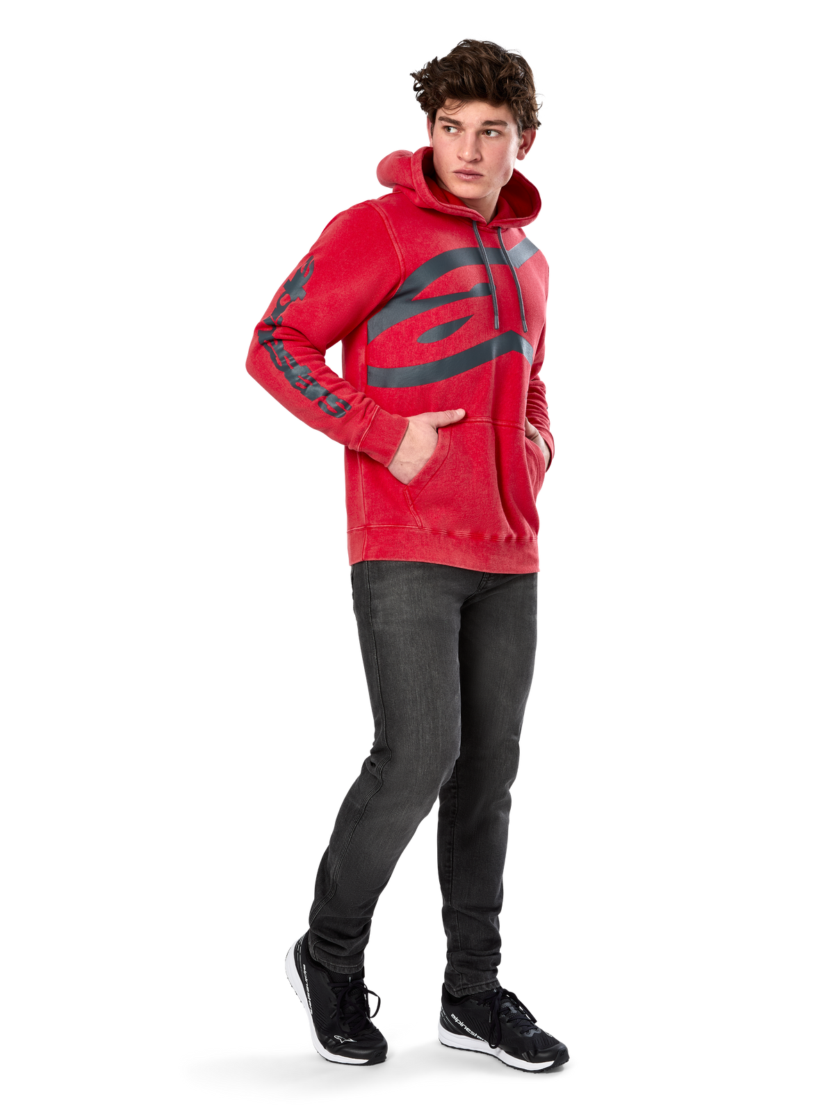Alpinestars Unbound Hoodie, röd plaggfärgad fleecehoodie i pullover-modell, extra stor mörk logotyp på bröstet och ordmärke på ärmarna, känguruficka, huva med dragsko, bärs av modell med mörkgrå jeans och svarta sneakers, avslappnad motorsportinspirerad streetwear