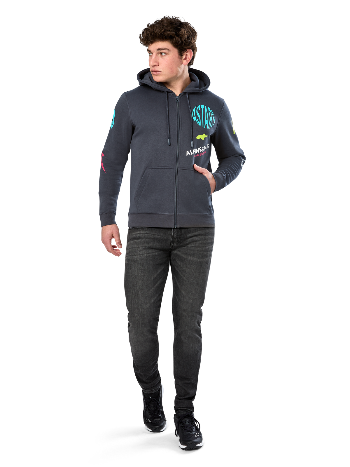 Alpinestars Variegate Hoodie, fleecehoodie med dragkedja framtill, gunmetal-grå, bärs av modell med svarta jeans och sneakers, med turkos 'ASTARS'-märkning och färgglad logografik på ärmar och bröst, avslappnad motorsportinspirerad streetwear