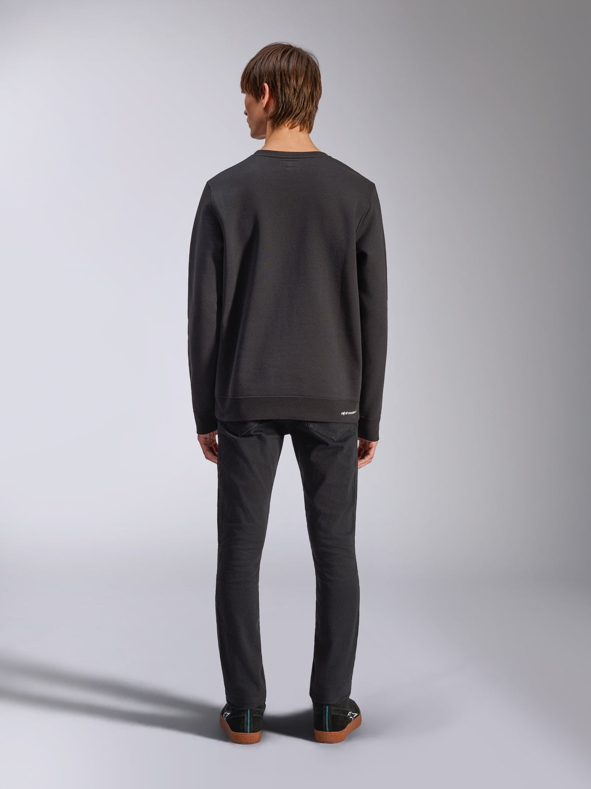 Alpinestars Positive Crew, svart sweatshirt, sedd bakifrån på manlig modell, bärs med svarta jeans och vardagliga sneakers, med ribbade muddar och fåll, motorsport-inspirerad streetwear