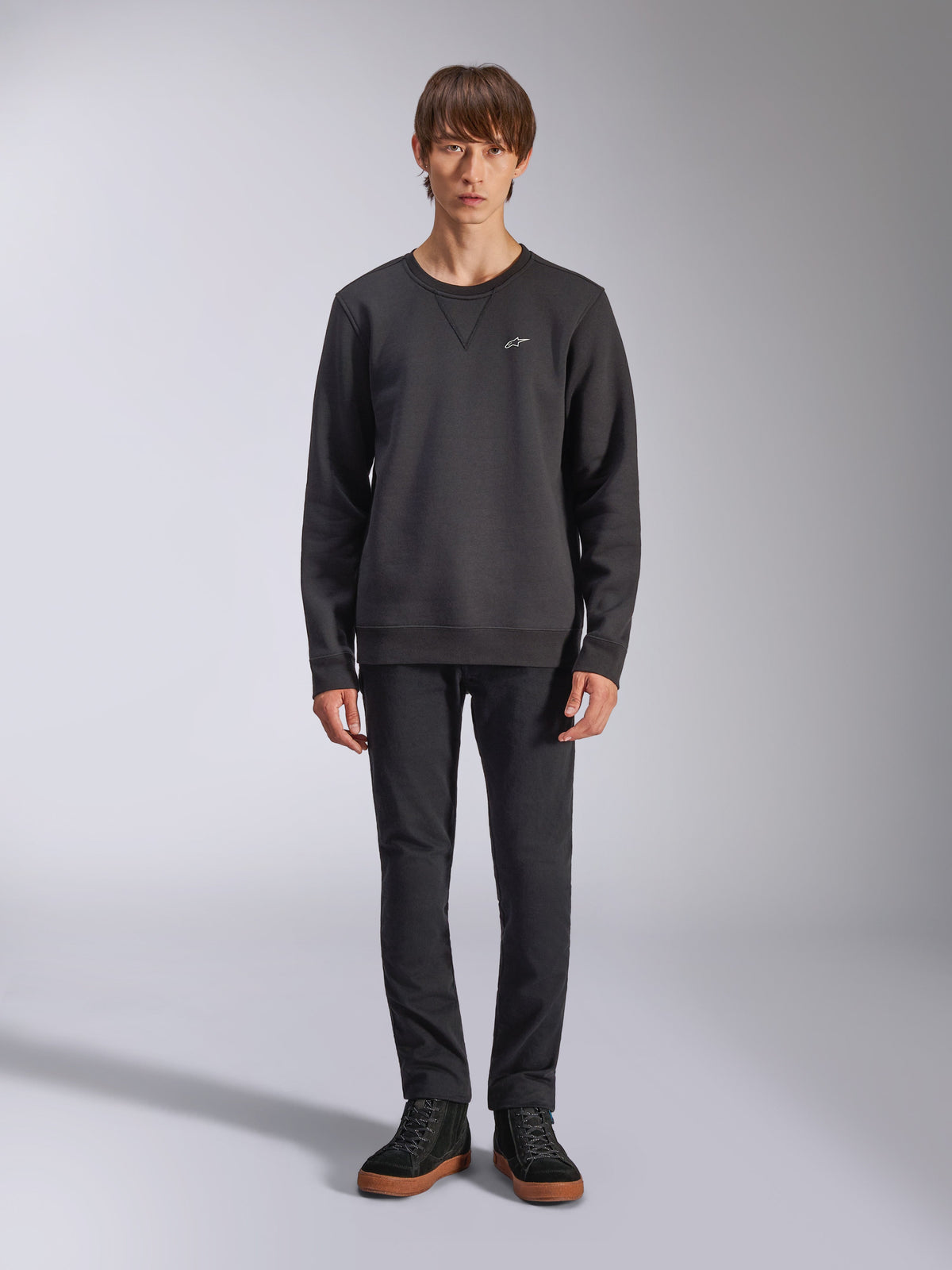 Alpinestars Positive Crew sweatshirt, avslappnad svart sweatshirt med rund hals, liten vit logotyp på bröstet, V-söm vid halsringningen, helkroppsbild i studio med modell som bär svarta byxor och sneakers, motorsportinspirerade livsstilskläder