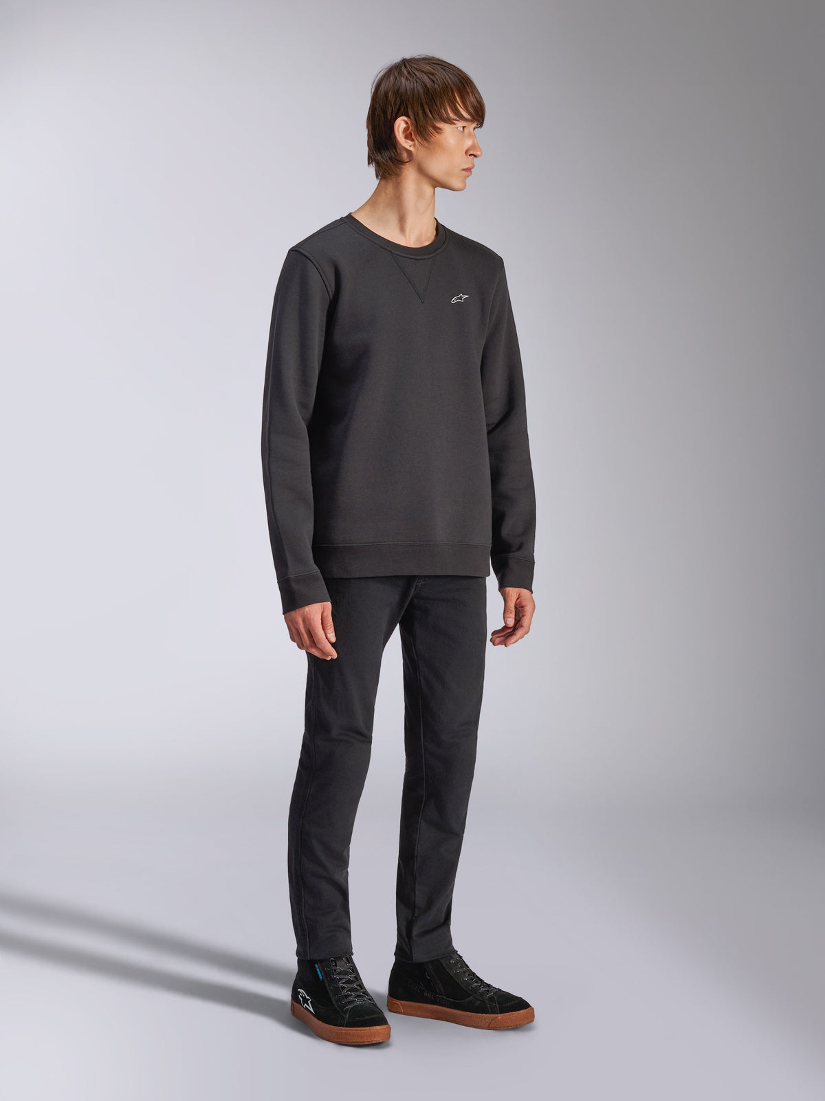 Alpinestars Positive Crew, svart sweatshirt, liten vit Alpinestars-logotyp på bröstet, ribbad krage med V-sömsdetalj, ribbade muddar och fåll, bärs av en modell med svarta byxor och sneakers, avslappnad motorsport-inspirerad streetwear
