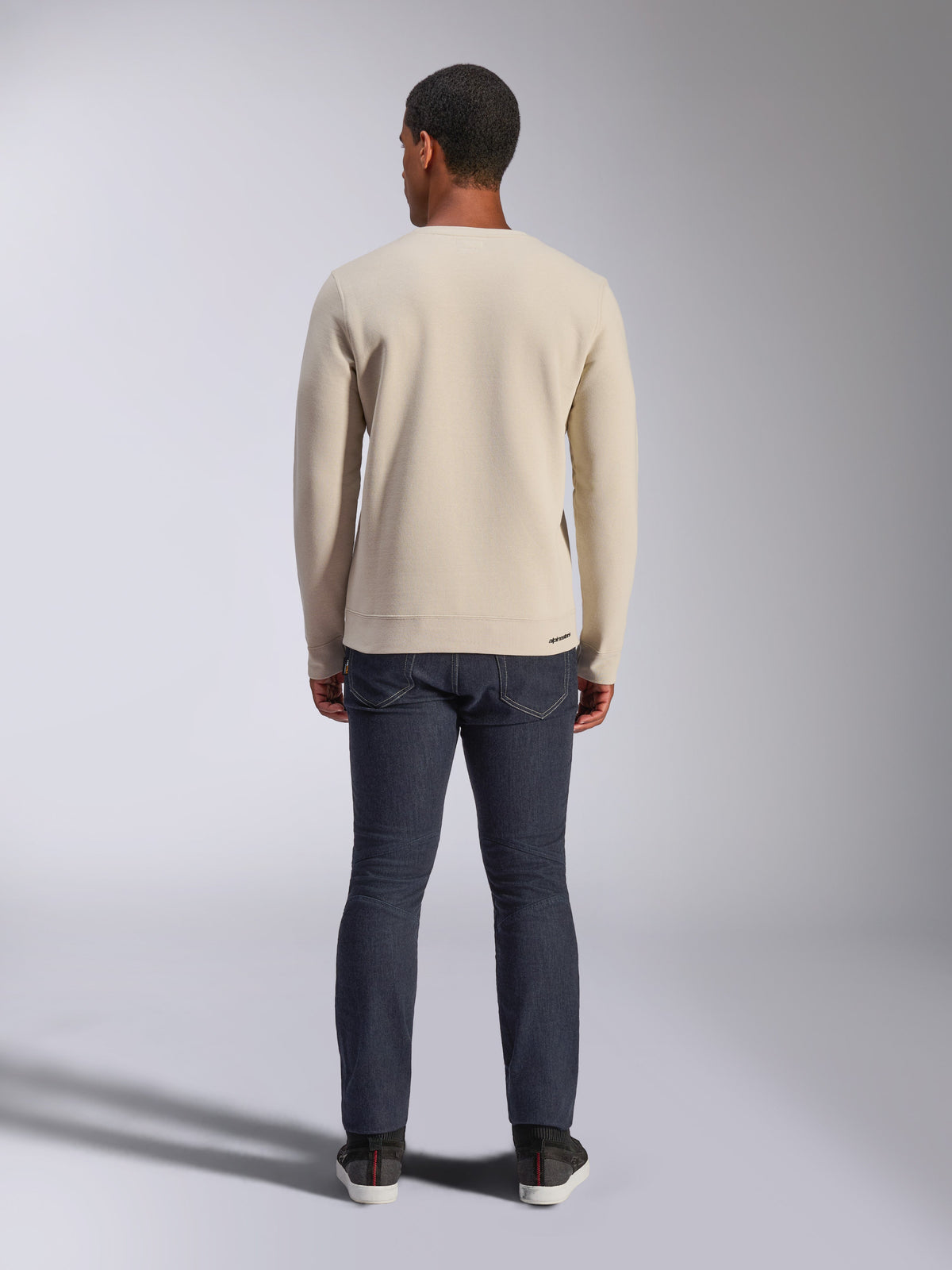Alpinestars Positive Crew Sweatshirt, Off White, baksida av en man som bär en beige sweatshirt med rund halsringning, ribbad fåll och muddar, kombinerat med mörka jeans, avslappnat motorsportinspirerat streetwear.