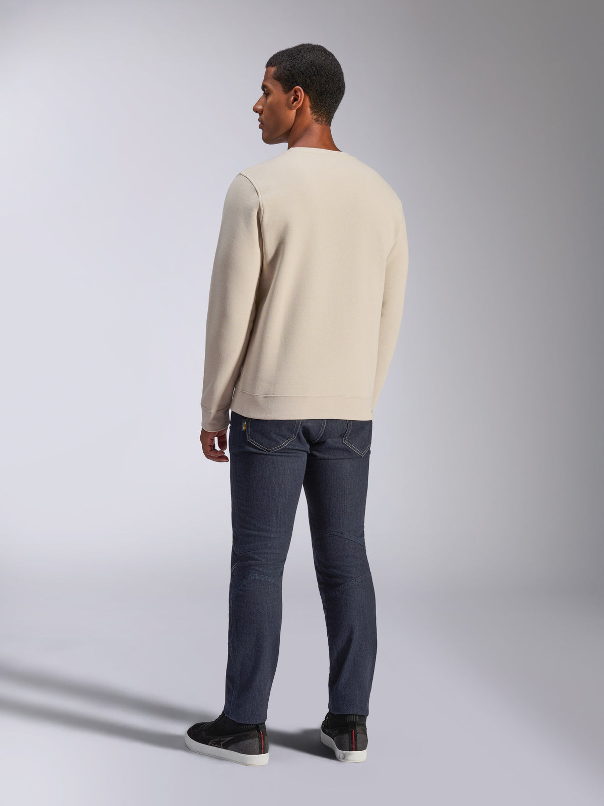 Alpinestars Positive Crew, Off White sweatshirt med rund halsringning, sedd bakifrån på modell, kombinerad med mörka denimjeans och svarta sneakers, avslappnad motorsport-streetwear, med ribbade muddar och fåll