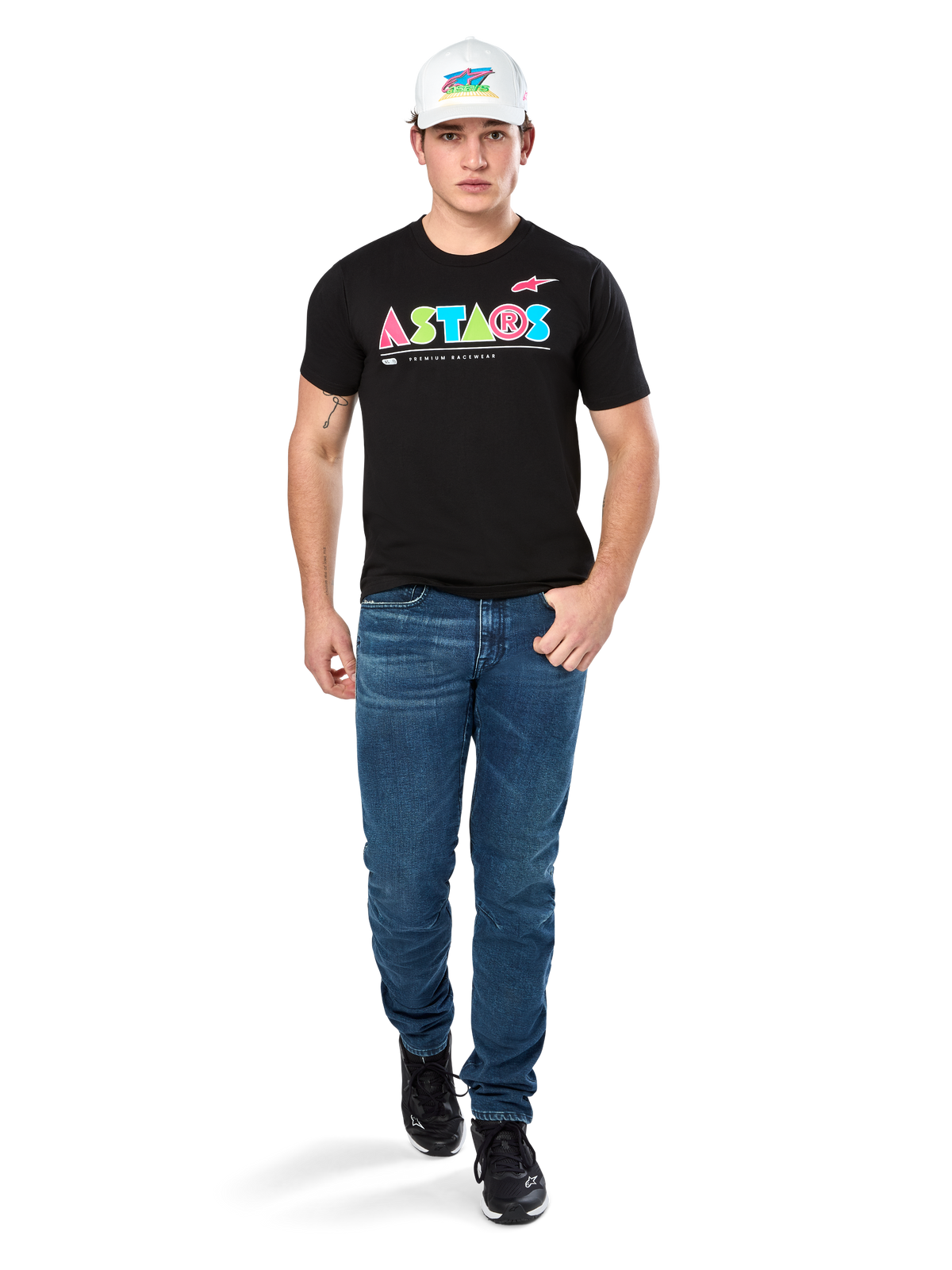 Male model wearing Alpinestars Cereal CSF Tee, Black Short Sleeve T-Shirt with colorful geometric ASTARS logo, paired with blue jeans and white cap, casual motorsport-inspired streetwear.