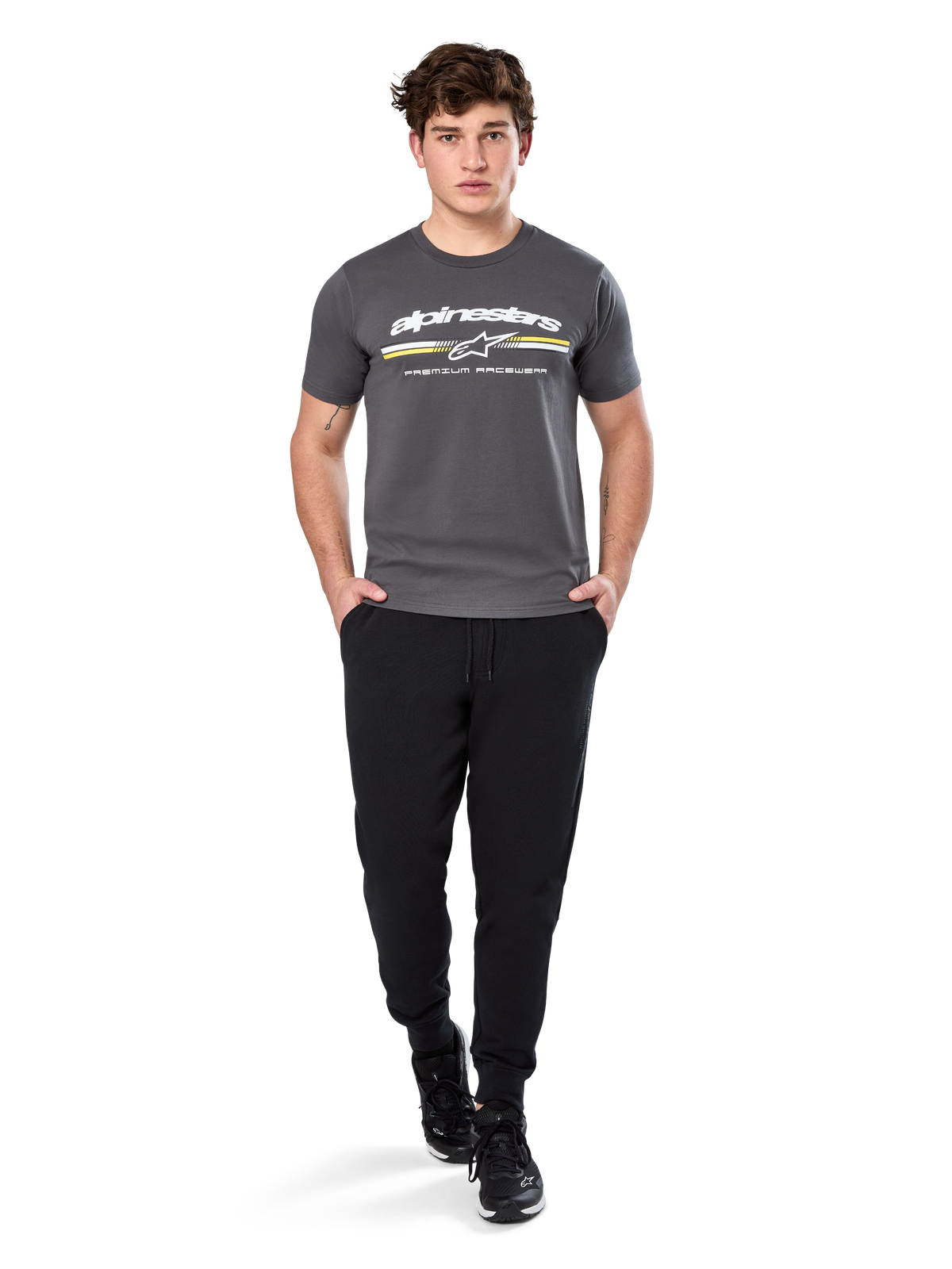 Man i Alpinestars Prevail Csf Tee, kortärmad t-shirt, mörkgrå, med vit Alpinestars-logotyp och trefärgad rand med gula detaljer, matchad med svarta joggers och sneakers, avslappnad motorsport-streetwear