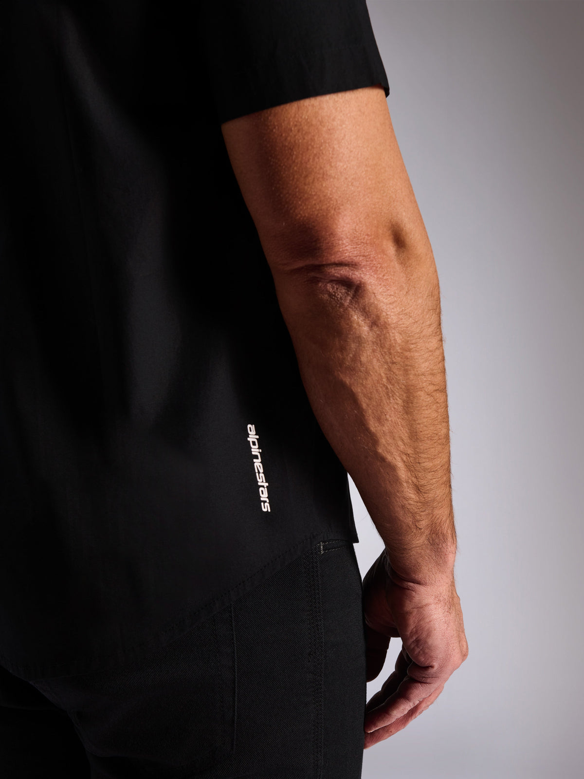 Alpinestars Strive Woven skjorta, svart kortärmad skjorta, vertikal vit Alpinestars-logotyp på sidan, närbildsdetalj av nederkanten och varumärkeslogotyp, casual motorsport-streetwear