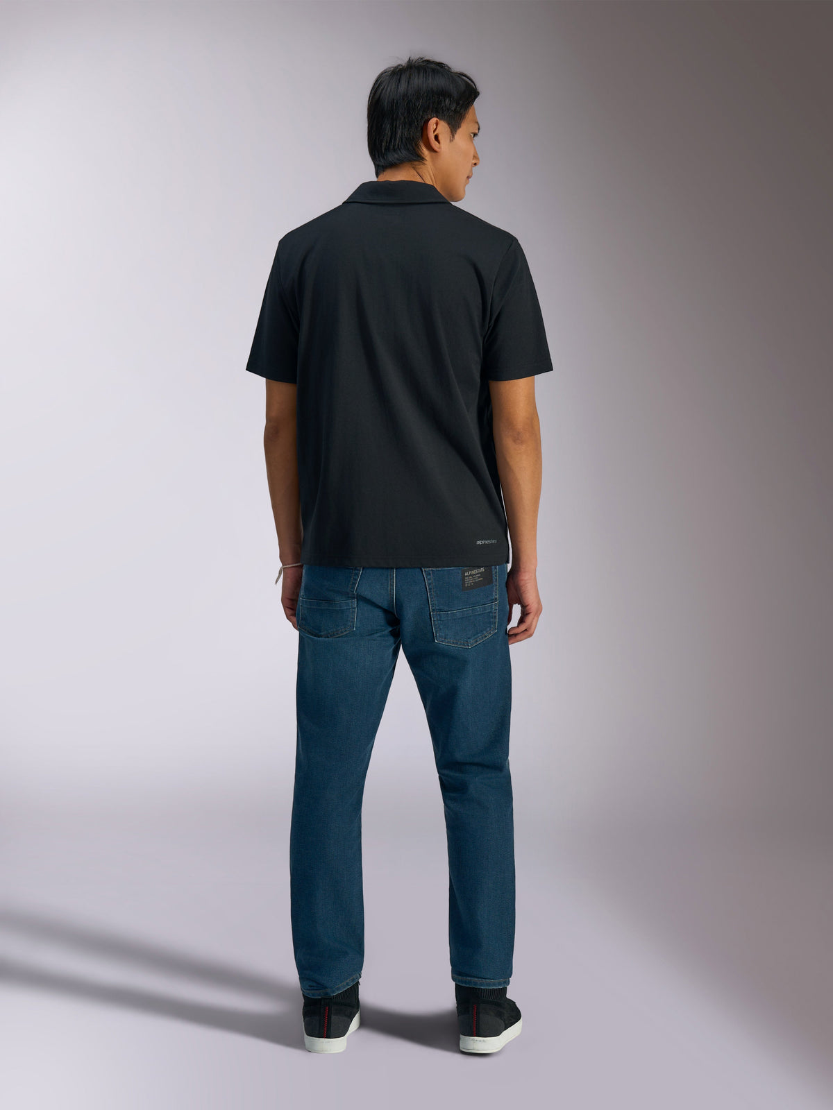Alpinestars Impetus Sport Polo, svart, baksida av kortärmad pikétröja med Alpinestars-logotyp på nedre ryggen, bärs med blå jeans, avslappnade motorsportinspirerade sportkläder