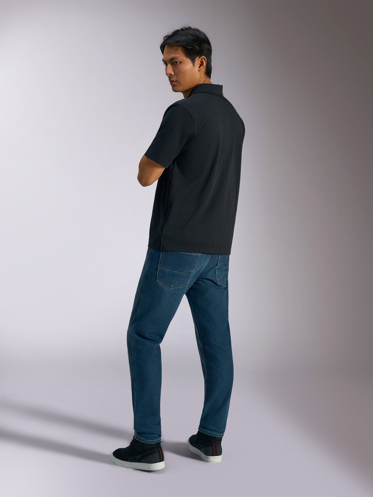 Alpinestars Impetus Sport Polo, svart, kortärmad pikétröja med mock mesh-textur och knappslå med tre knappar, bärs av en modell med blå jeans och svarta sneakers, vardaglig motorsportinspirerad streetwear