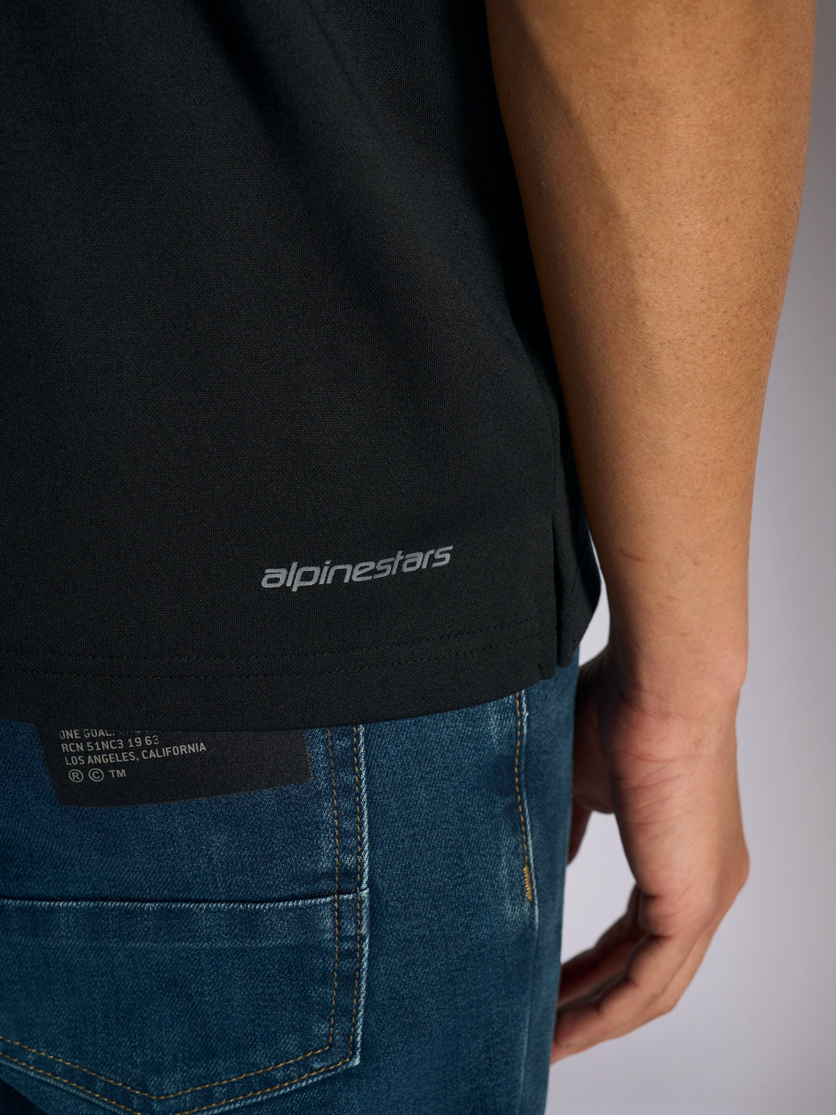 Alpinestars Impetus Sport Polo, detaljvy av den nedre fållen och sidoslitsen, svart med grått Alpinestars-ordmärke, vardagliga motorsportinspirerade sportkläder