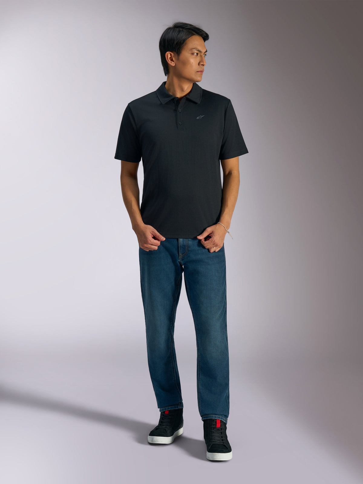 Alpinestars Impetus Sport Polo, svart kortärmad pikétröja, har en knappslå med tre knappar och grå Astars-logotyp på bröstet, bärs av en man med blå jeans och svarta high-top sneakers, avslappnade motorsport-inspirerade sportkläder