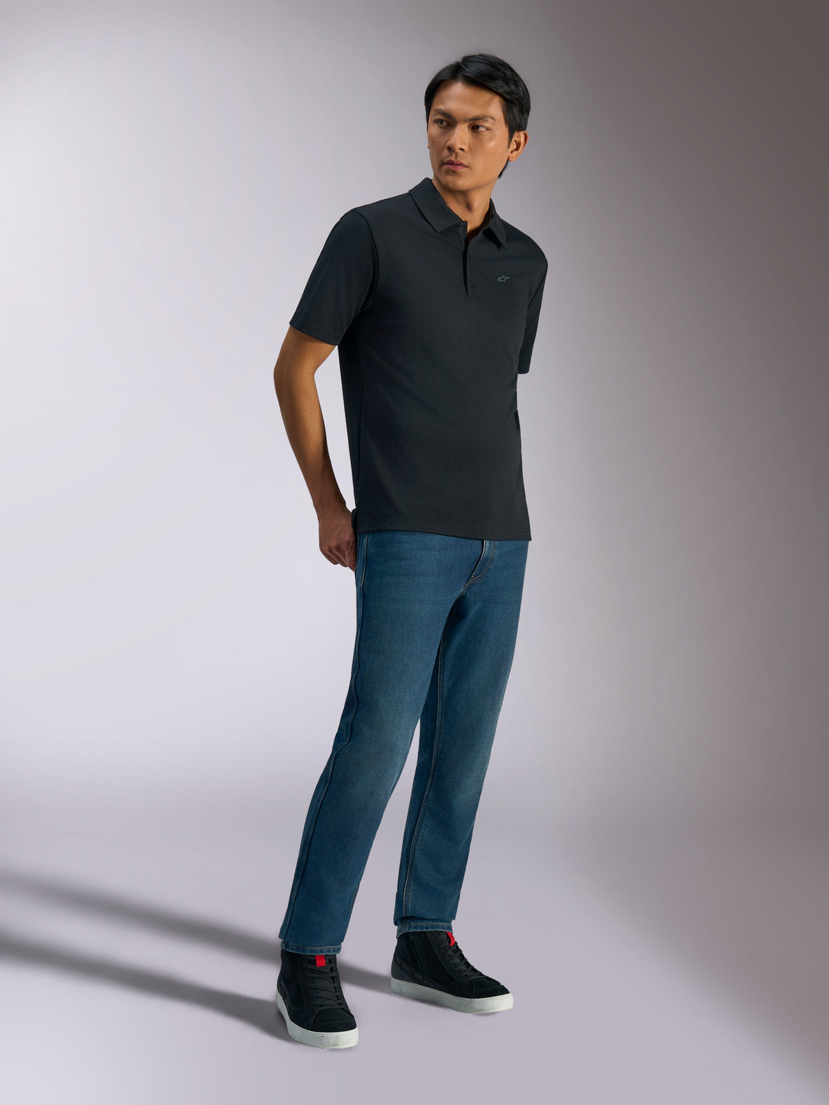 Alpinestars Impetus Sport Pikétröja, kortärmad pikétröja, svart, med en diskret logotyp på bröstet och knappslå med tre knappar, bärs av en modell med blå jeans och svarta sneakers, vardagliga motorsportinspirerade livsstilskläder