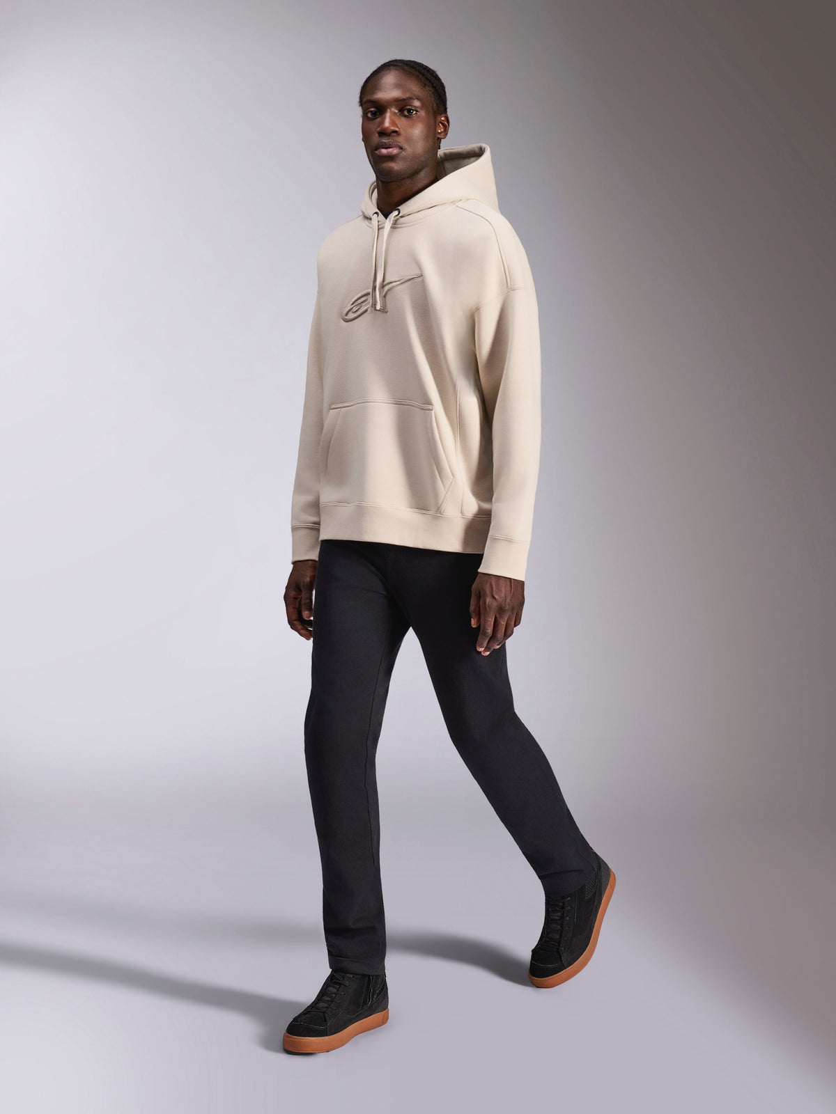 Alpinestars Emergent Oversized Hoodie, vardaglig streetwear, sandfärgad off-white, ton-i-ton 3D-broderad logotyp, bärs av modell med svarta byxor och svarta sneakers, avslappnad oversized passform med känguruficka och huva med dragsnöre