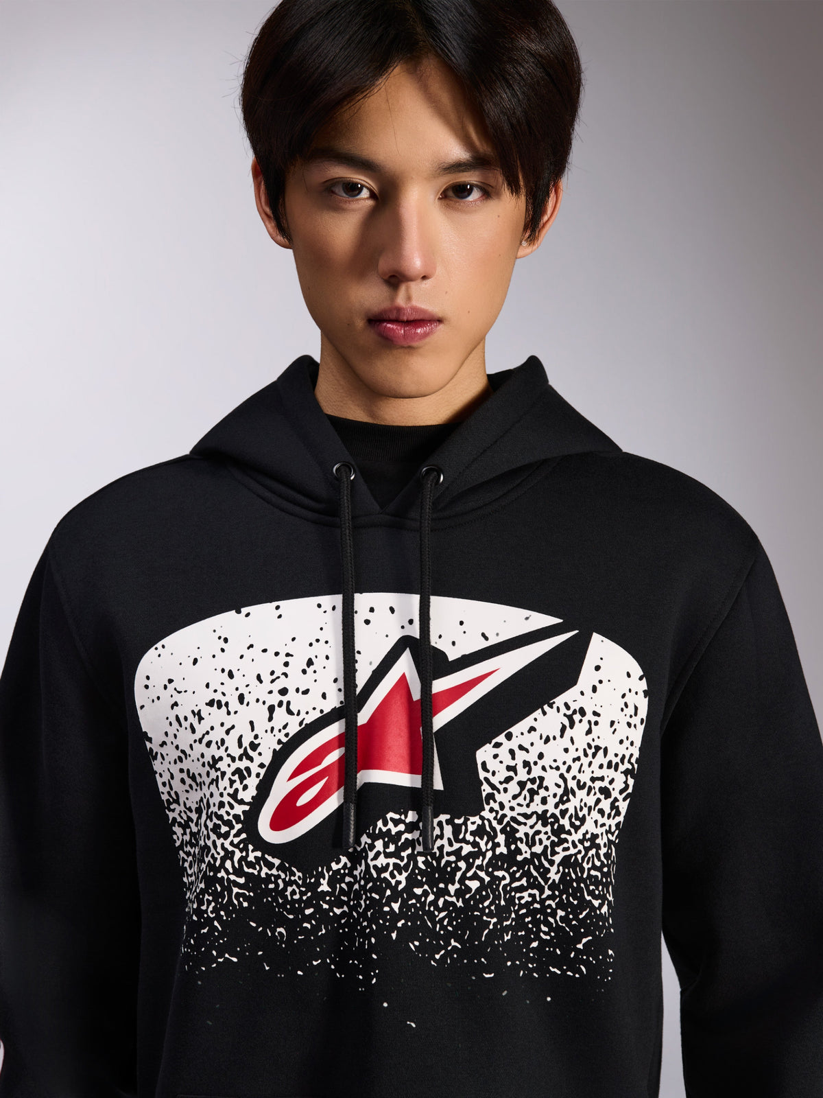 Alpinestars Obsessed Hoodie, svart pullover i fleece, med en röd och vit Astars-logotyp inuti en vit prickig MX-graderingsgrafik på bröstet, bärs av en modell, avslappnat motorsport-streetwear med justerbar huva med dragsnöre