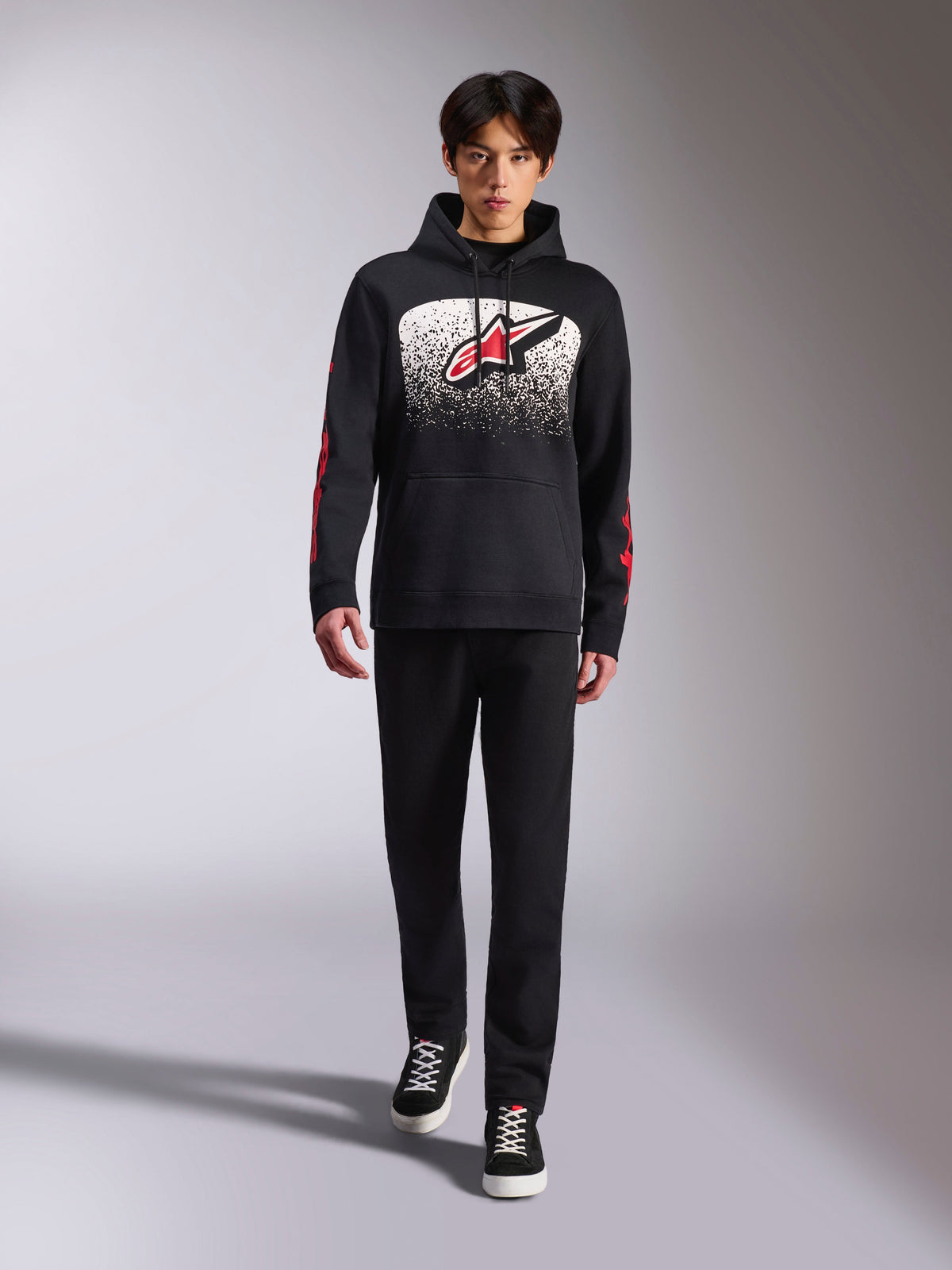 Alpinestars Obsessed Hoodie, svart huvtröja, stor vit och röd Astars-logotyp med MX-gradering, röda ordlogotyper på ärmarna, bärs av modell i en helsvart avslappnad outfit, motorsportinspirerade livsstilskläder