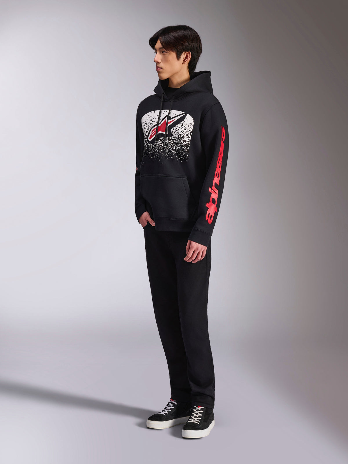 Alpinestars Obsessed Hoodie, svart huvtröja i fleece, med en stor vit spräcklig grafik på bröstet, röd Astars-logotyp och röd text på ärmarna, bärs av en modell med svarta byxor och sneakers, avslappnad motorsport-streetwear