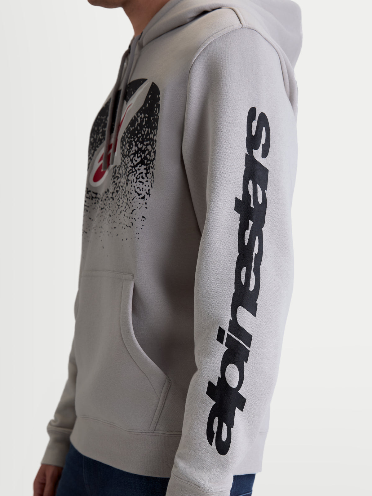 Alpinestars Obsessed huvtröja, motorsportinspirerad fleecehuvtröja, unisex, cementgrå och svart, sidovy på modell som framhäver vertikal Alpinestars-profilering på ärmen och MX-inspirerad logotyp på bröstet, tillverkad i bomull och återvunnen polyester.