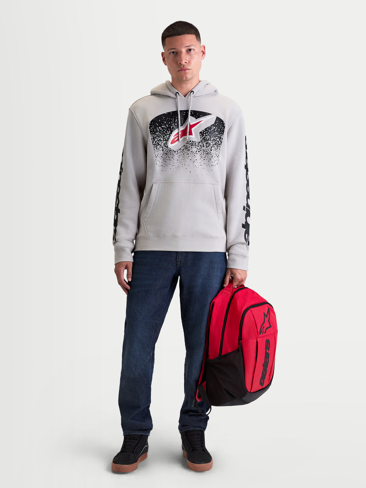 Alpinestars Obsessed huvtröja, sportig herrhuvtröja i fleece, grå med röd och svart MX-inspirerad grafik på bröstet och ärmarna. Denna bekväma lifestyle-huvtröja visas på en manlig modell som håller en röd Alpinestars ryggsäck.