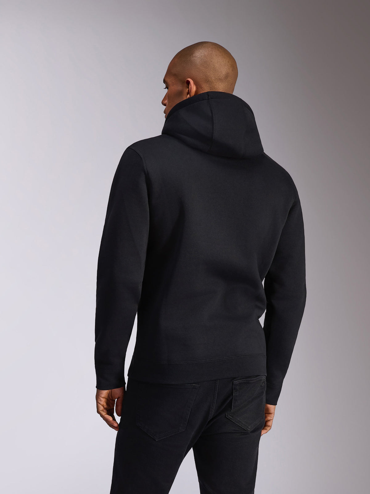 Alpinestars Ellipsoid Hoodie, svart, baksida av en person som bär en huvtröja i fleece, långa ärmar med ribbade muddar och fåll, stylad med svarta jeans, vardagliga motorsportinspirerade sportkläder