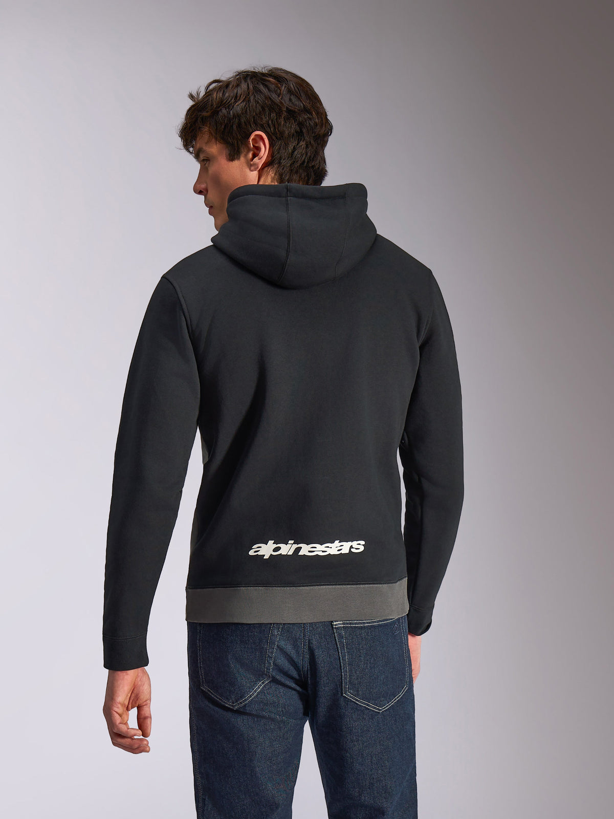 Alpinestars Parallel Hoodie, avslappnad huvtröja, svart och gunmetal, vy bakifrån med en vit Alpinestars-logotyp på nedre delen av ryggen, färgblockerad design med grå fåll, bärs med mörka denimjeans
