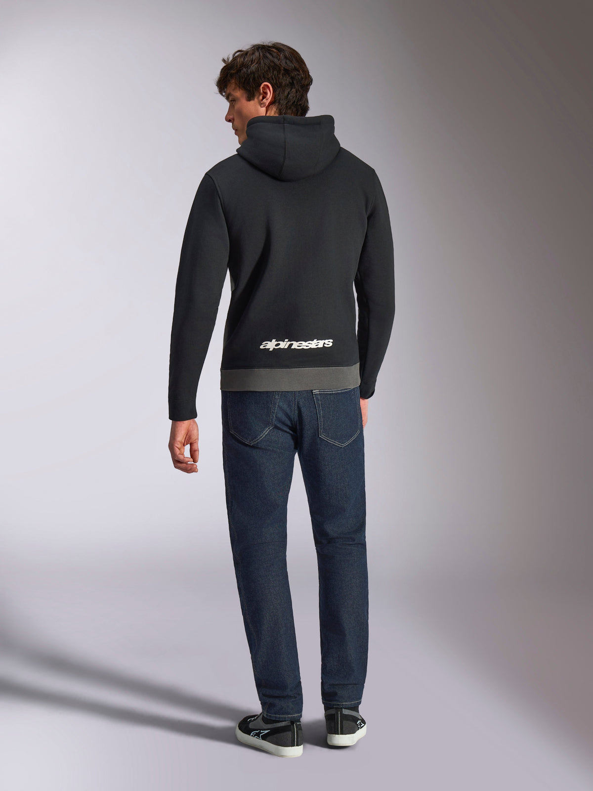 Alpinestars Parallel Huvtröja, baksida av en man i en svart och gunmetal färgblockad pullover-fleece, vit Alpinestars-logotyp på nedre delen av ryggen, matchad med mörkblå jeans, avslappnad motorsport-inspirerad streetwear