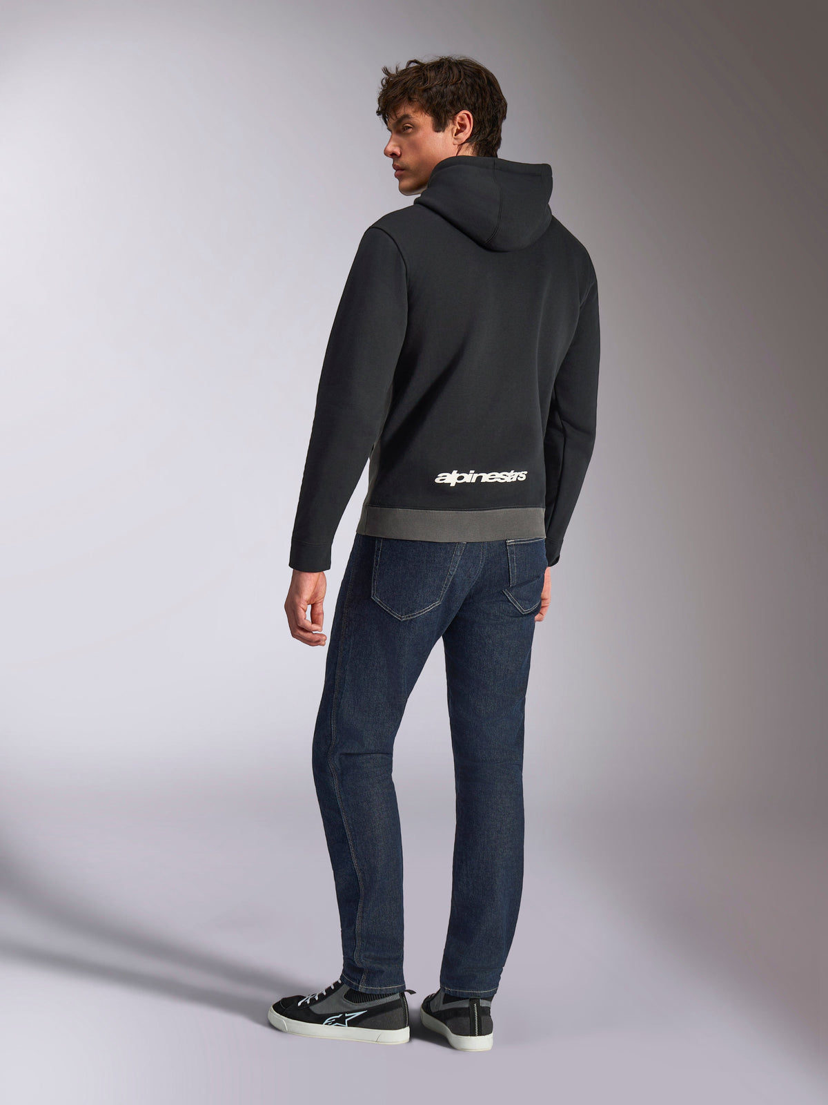 Alpinestars Parallel Hoodie, vy bakifrån av modell som bär den svarta och gunmetal-gråa huvtröjan, med vit Alpinestars-logotyp på nedre delen av ryggen, matchat med mörkblå jeans, avslappnad motorsport-inspirerad streetwear