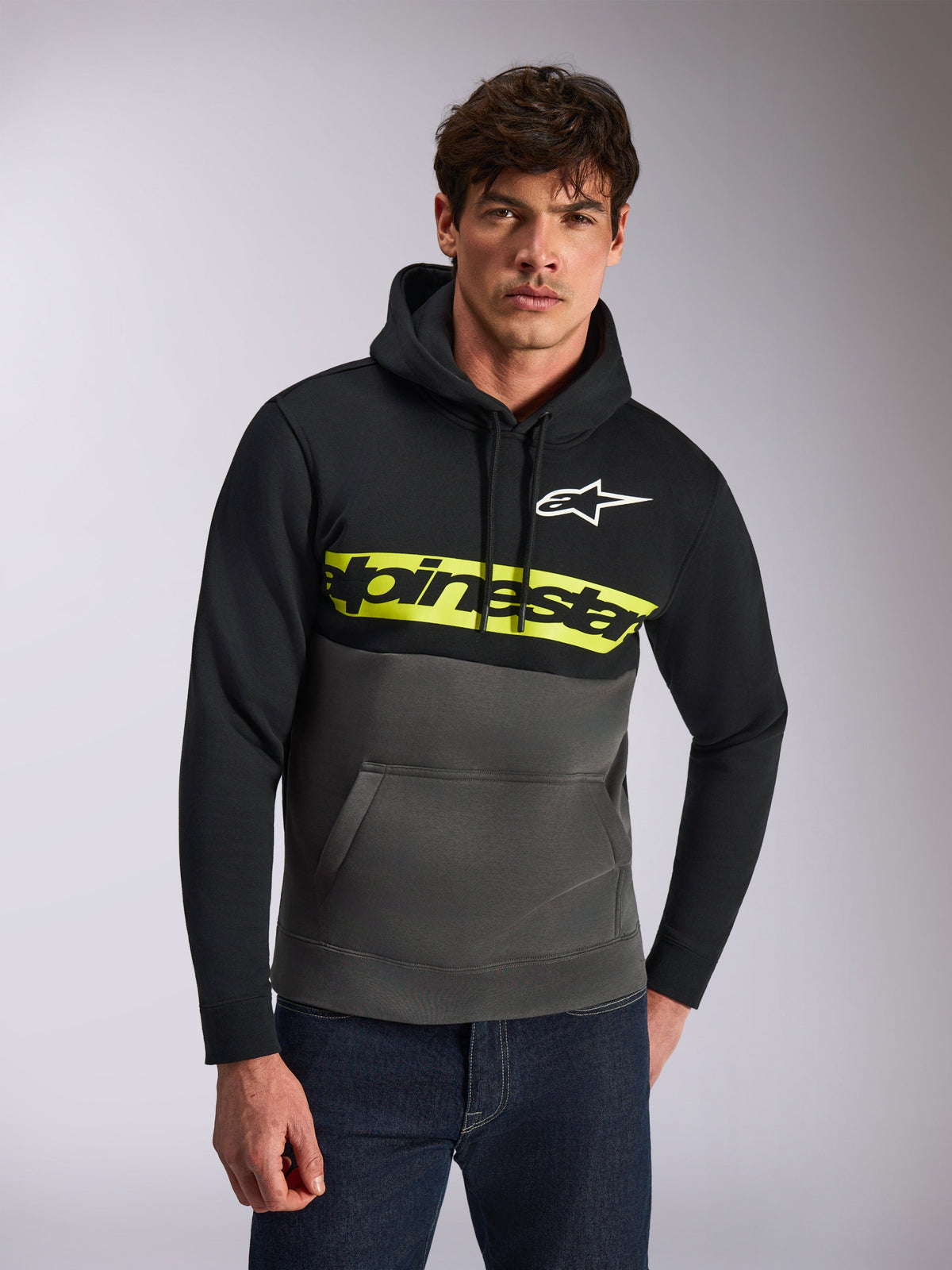 Alpinestars Parallel Hoodie, vardaglig streetwear, färgblockerad design i svart och gunmetal-grå, gult bröstband med Alpinestars ordlogotyp, vit Astars-logotyp, bärs av en modell i blå jeans, modern motorsportinspirerad design