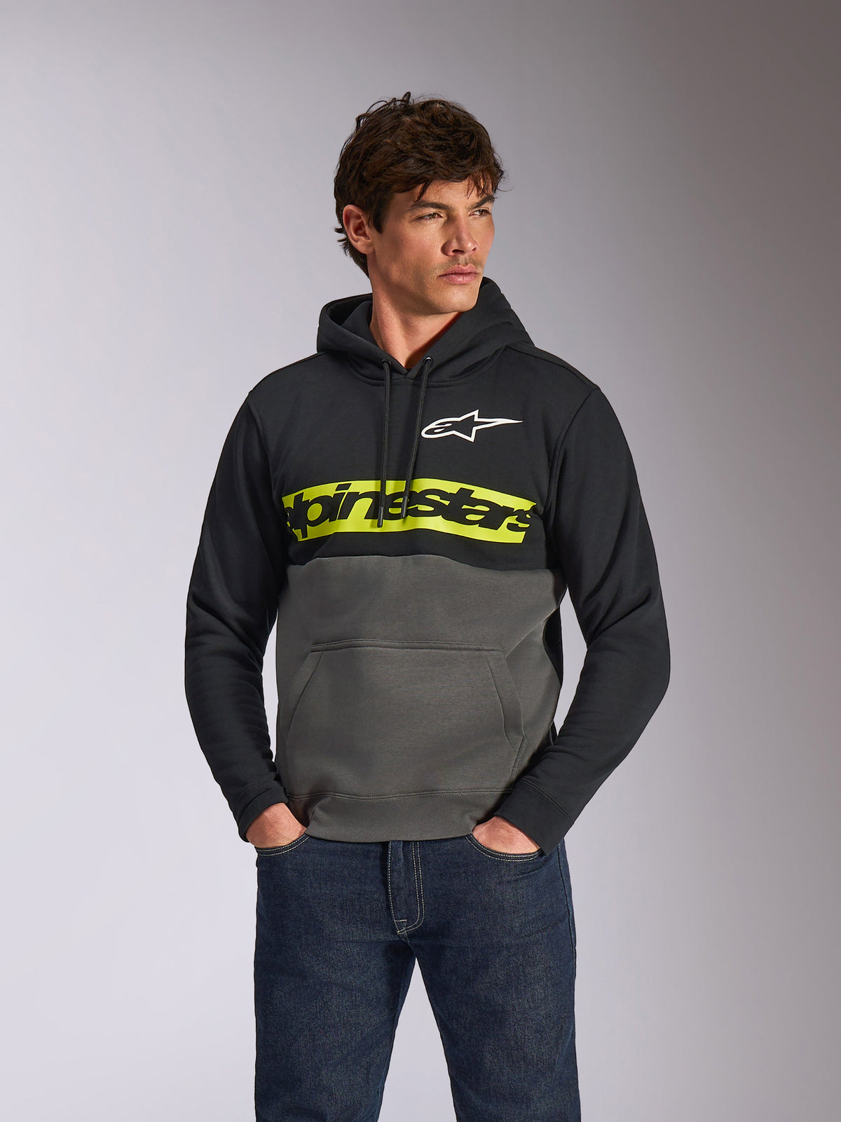 Alpinestars Parallel Hoodie, avslappnad färgblockerad huvtröja i fleece, svart och gunmetal med ett klart gult bröstband, Alpinestars ordlogga och vit stjärnlogotyp, bärs av en manlig modell med mörkblå jeans