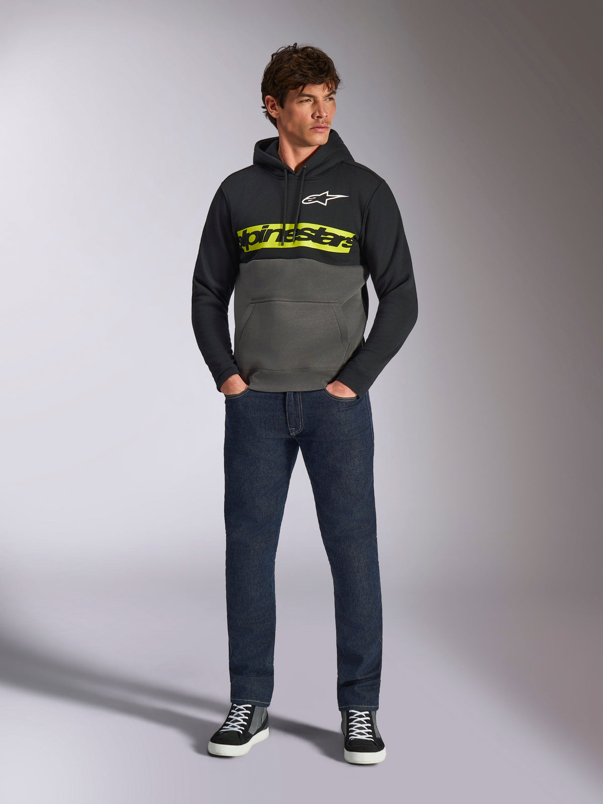 Alpinestars Parallel Hoodie, färgblockerad huvtröja, svart och gunmetal-grå, klargul Alpinestars-ordlogotyp och vit stjärnlogotyp på bröstet, bärs av en manlig modell med mörkblå jeans och svarta sneakers, avslappnad motorsport-streetwear