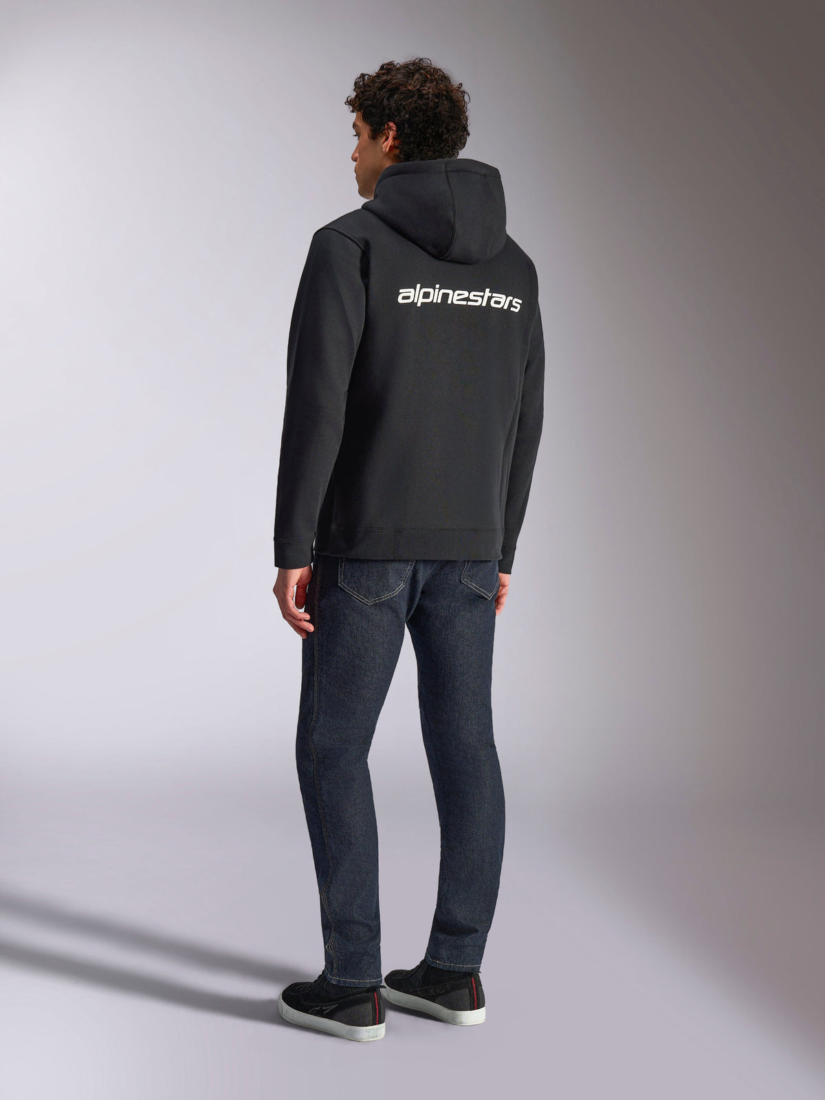 Alpinestars Transverse Huvtröja, svart fleece med dragkedja fram, vy bakifrån med en vit Alpinestars-ordlogotyp över axlarna, bärs med mörkblå jeans och svarta sneakers, avslappnat motorsportinspirerat streetwear