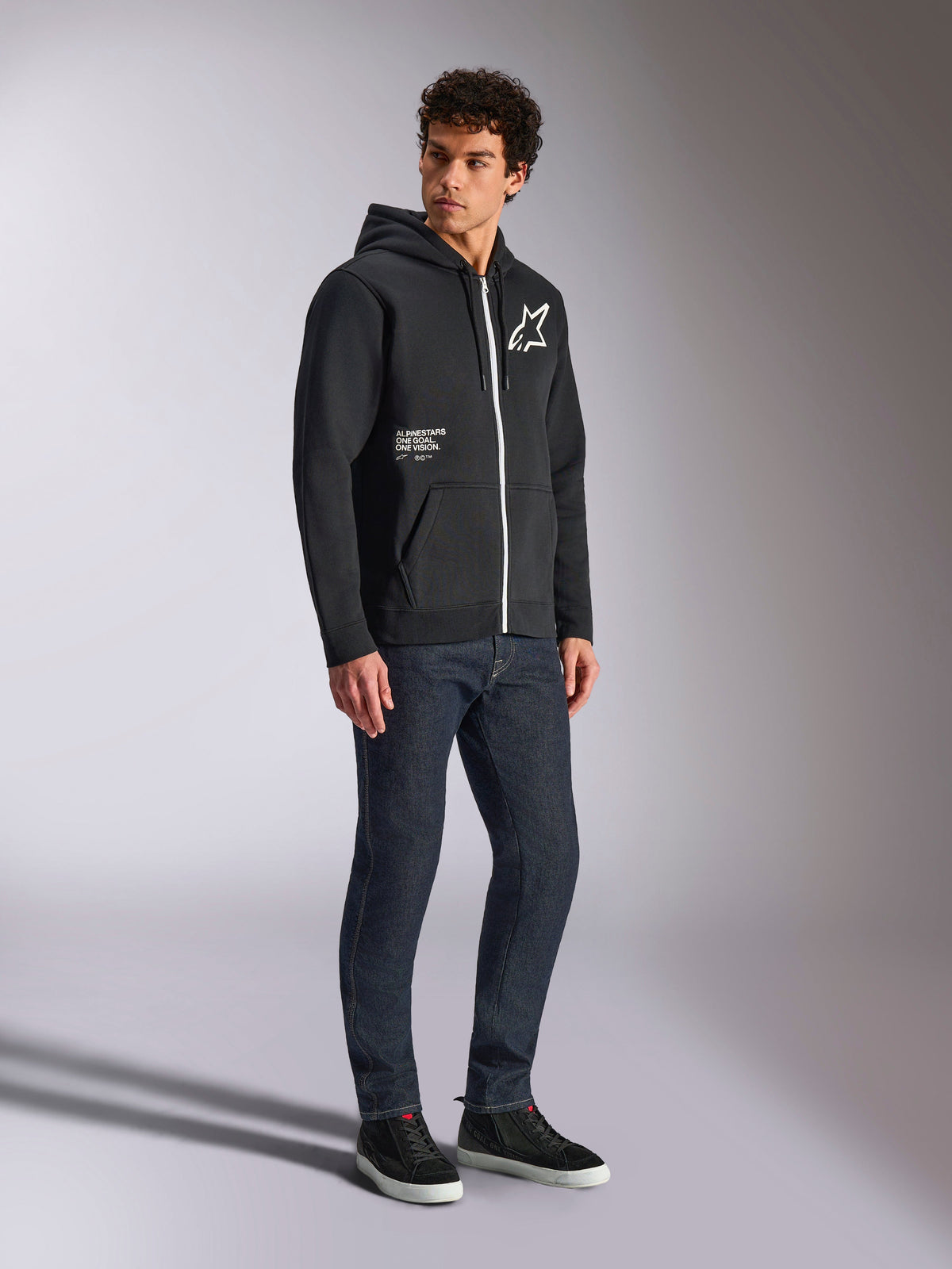 Alpinestars Transverse Hoodie, svart huvtröja med dragkedja och en kontrasterande vit dragkedja, med en vit stjärnlogotyp på bröstet och 'One Goal'-branding på fickan, bärs av en modell med mörka jeans och svarta sneakers, avslappnad motorsportinspirerad streetwear.