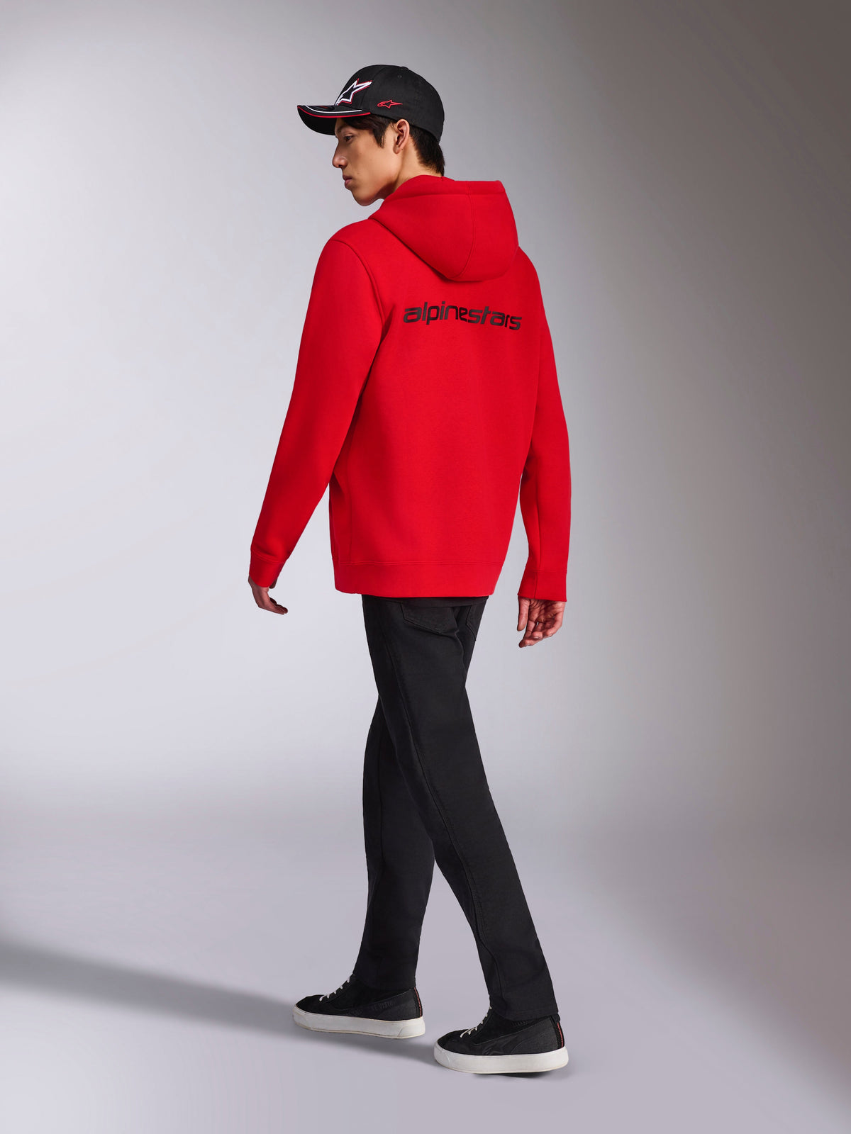 Alpinestars Transverse Hoodie, röd fleecehoodie med dragkedja, baksida med svart Alpinestars-logotyp, bärs av en modell med svarta byxor och en svart keps med logotyp, avslappnad motorsport-inspirerad streetwear