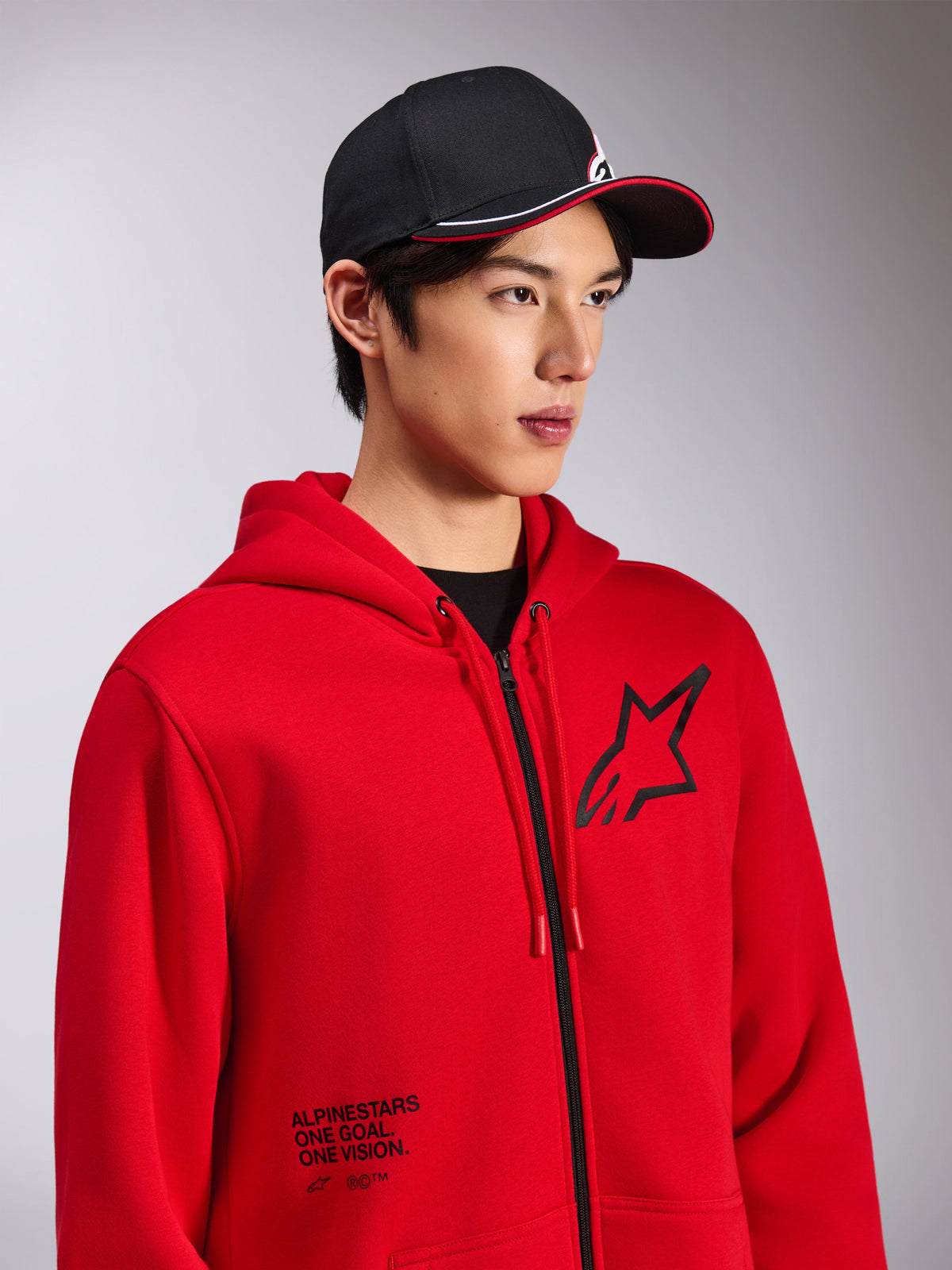 Alpinestars Transverse Hoodie, röd fleecehuvtröja med dragkedja fram, svart Astars-logotyp på bröstet och texten 'One Goal. One Vision.' nedtill på framsidan, casual motorsport-streetwear, bärs av en modell med en svart keps