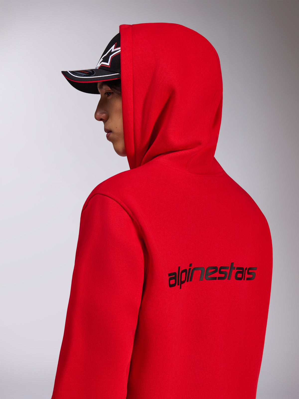 Alpinestars Transverse Hoodie, röd huvtröja i fleece, vy snett bakifrån som visar den svarta Alpinestars-ordlogotypen på ryggen, bärs med huvan uppe och en svart keps, avslappnad motorsportinspirerad streetwear