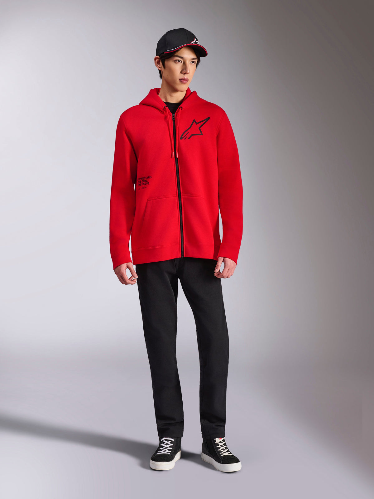 Alpinestars Transverse Hoodie, röd fleecehuvtröja med hel dragkedja, svart Astars-logotyp på bröstet och One Goal-tryck på sidan, bärs av manlig modell med svarta byxor och keps, avslappnad motorsport-inspirerad streetwear