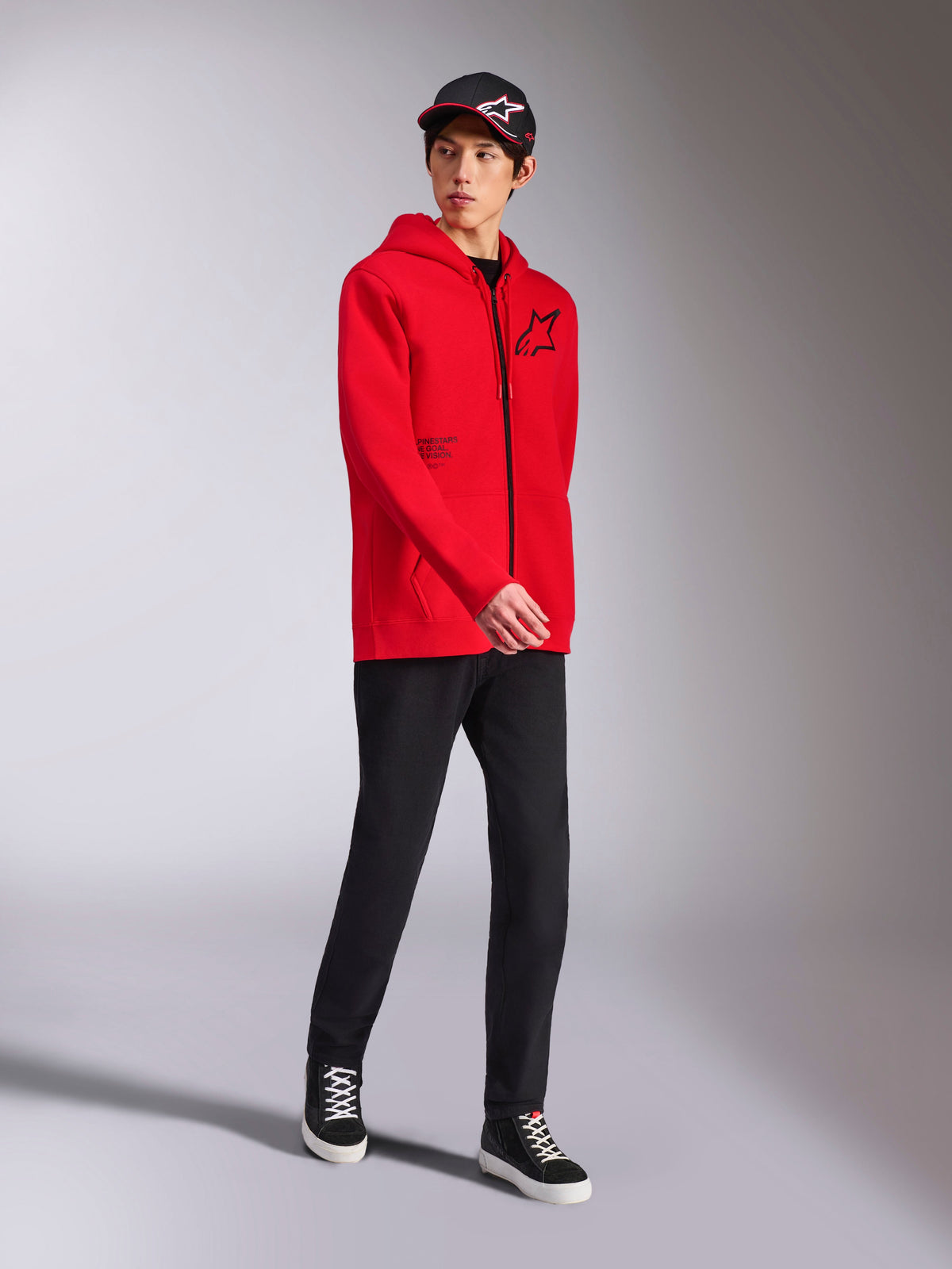 Alpinestars Transverse Hoodie i rött, manlig modell bär huvtröja i fleece med dragkedja, svart Astars-logotyp och texttryck, stylad med svarta byxor och keps, avslappnad motorsport-streetwear