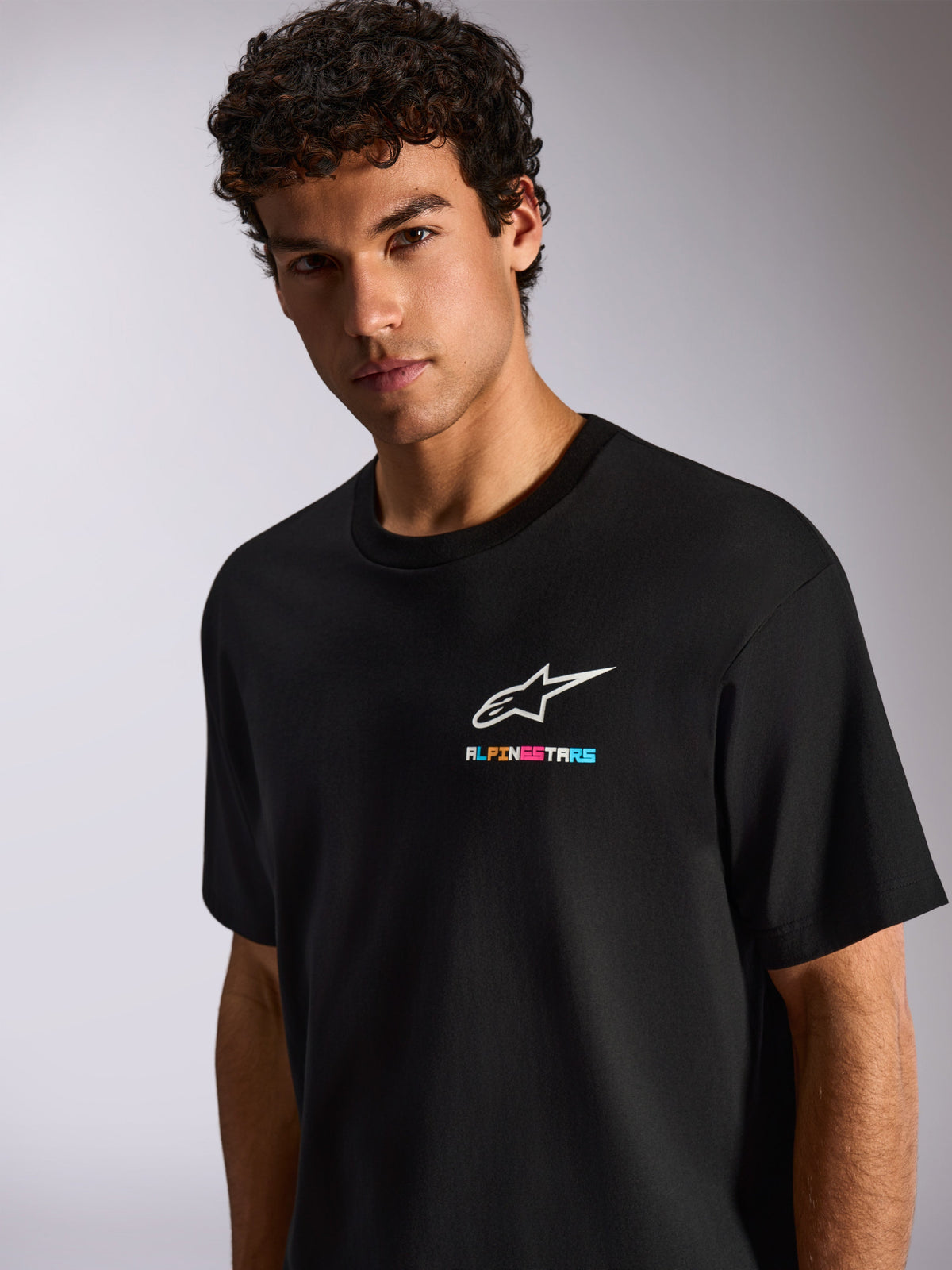 En man i Alpinestars Wild Oversized CSF Tee, svart kortärmad t-shirt med en vit Astars-logotyp och livfullt flerfärgat tryck på bröstet, avslappnad motorsport-streetwear