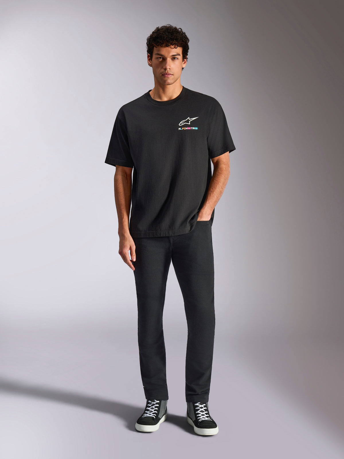 Alpinestars Wild Oversized CSF Tee, kortärmad t-shirt, svart, bärs av en modell med svarta byxor och sneakers, med en vit Astars-logotyp och livfull flerfärgad grafik, avslappnad motorsport-streetwear