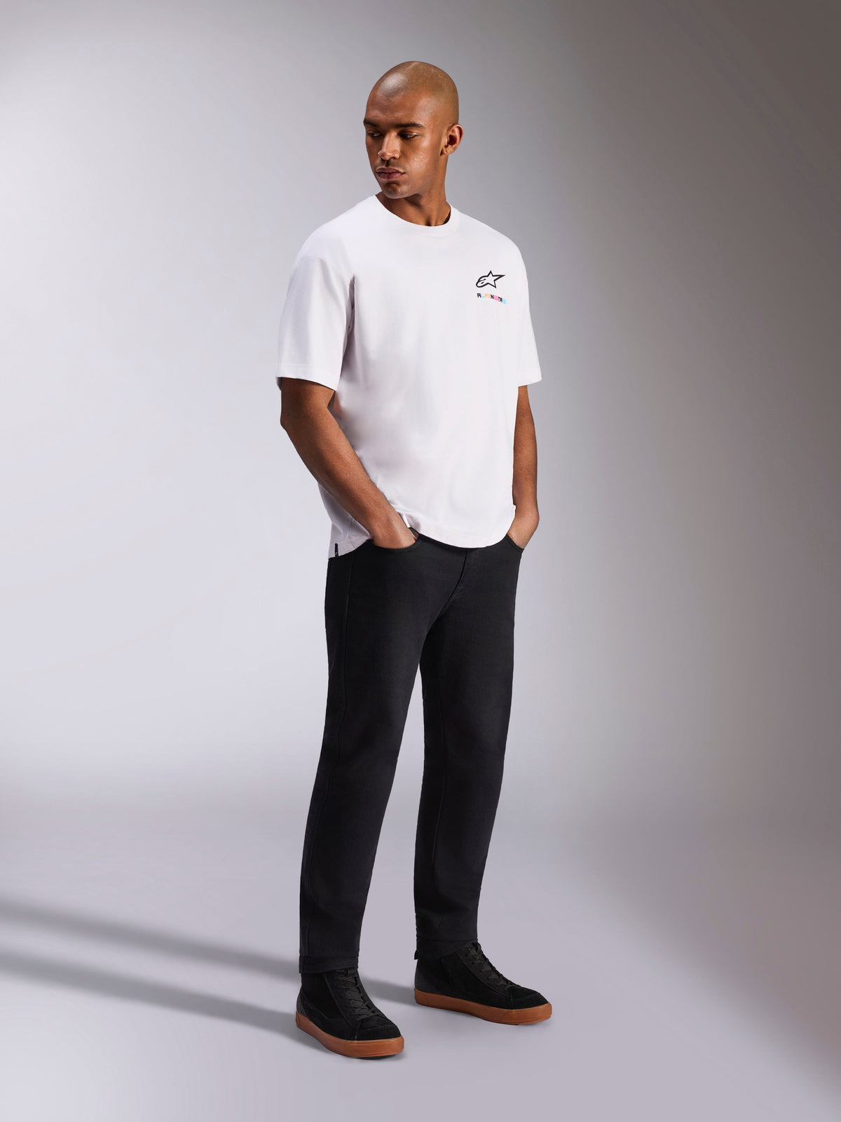 Alpinestars Wild Oversized CSF Tee, kortärmad t-shirt, vit, med en liten flerfärgad Astars-logotyp på bröstet, bärs av en modell med svarta byxor och svarta sneakers, avslappnat motorsportinspirerat streetwear