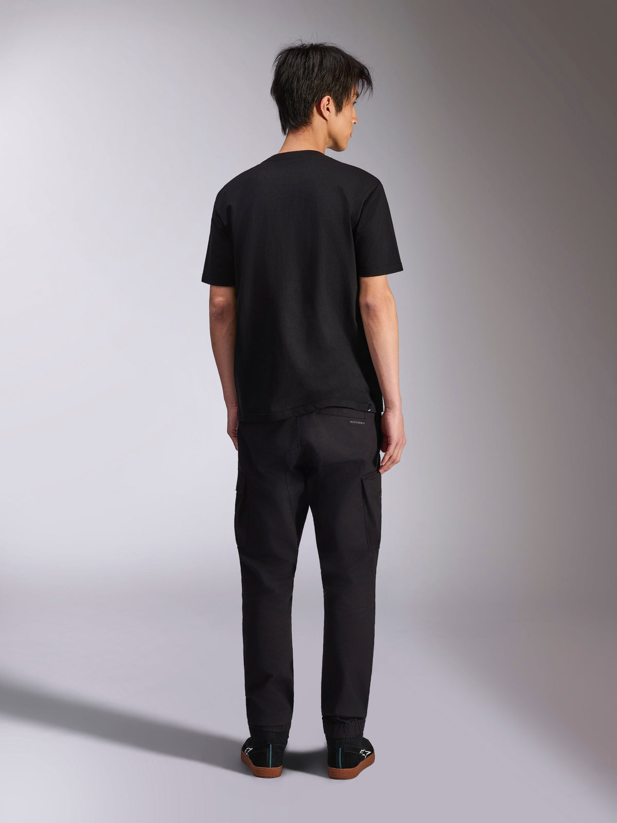 Alpinestars Noka CSF Tee, kortärmad t-shirt, svart, ryggvy av modell i svarta byxor och sneakers, avslappnad motorsport-streetwear-stil
