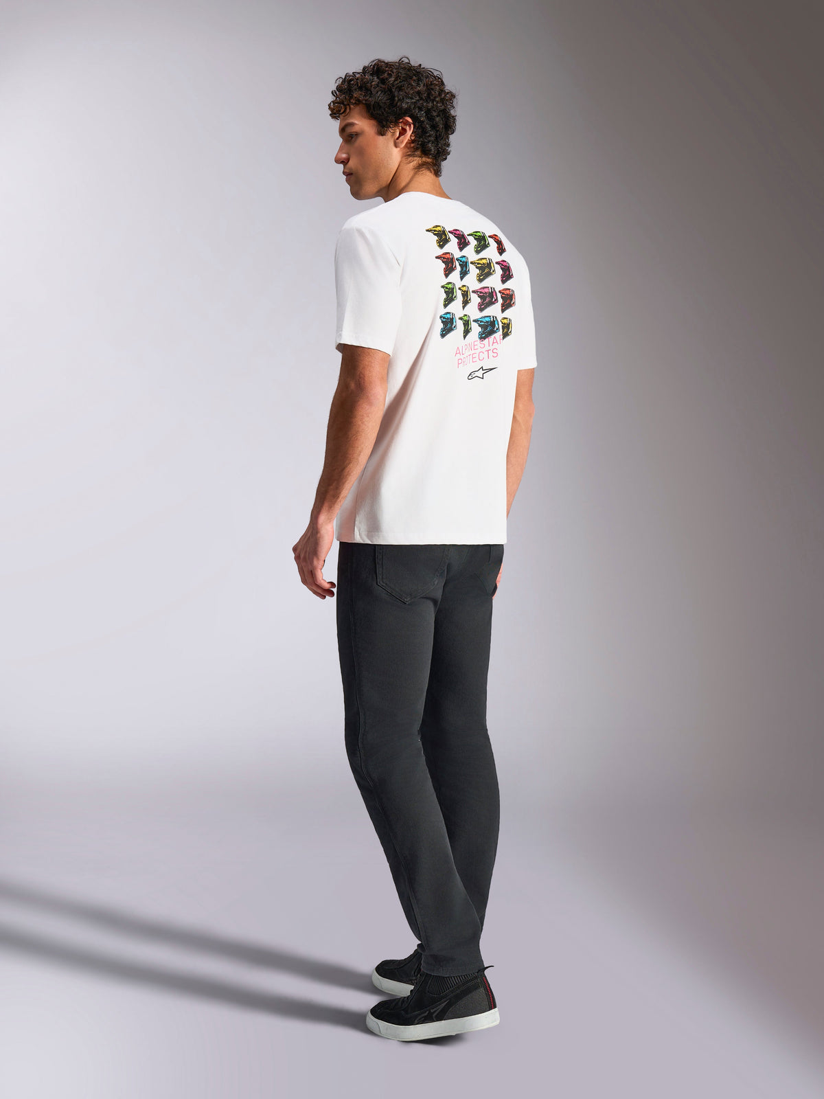 Alpinestars Pop Helmet CSF Tee, kortärmad t-shirt, vit, baksida på modell som visar färgglad M10-hjälmkonst, stylad med mörka byxor och svarta sneakers, avslappnad motorsportinspirerad streetwear