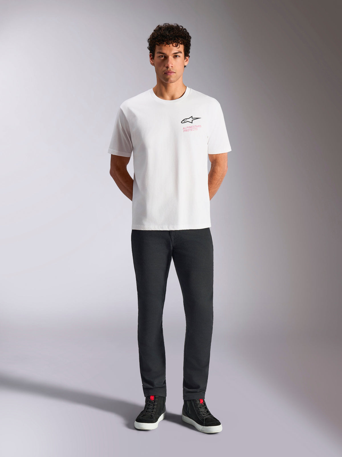 Alpinestars Pop Helmet CSF Tee, kortärmad t-shirt, vit, bärs av en modell med svarta byxor och svarta höga sneakers, Astars-logotyp på framsidan med rosa 'Alpinestars Protects'-text, avslappnat motorsport-streetwear