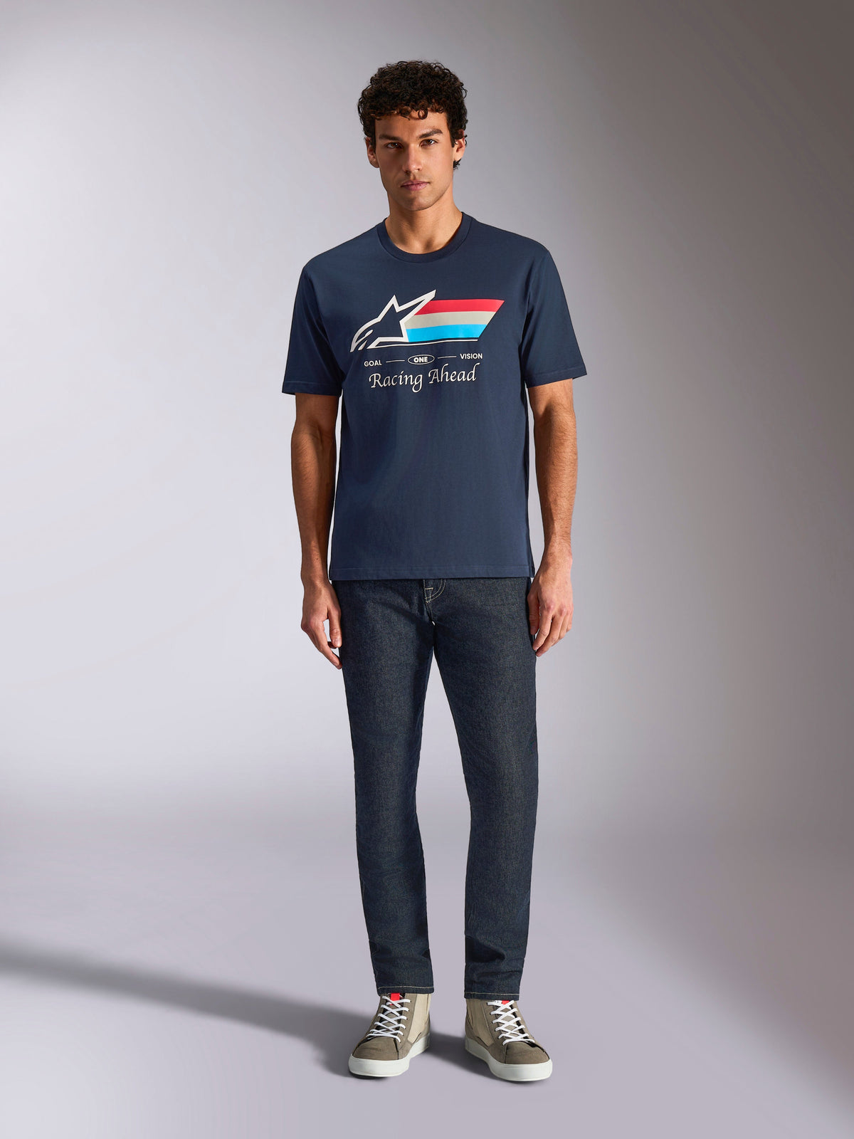 Alpinestars Trifecta CSF Tee, kortärmad t-shirt, marinblå, vit Astars-logotyp med röda, vita och ljusblå ränder och texten "Racing Ahead", bärs med mörka jeans och olivgröna sneakers, avslappnad motorsport-streetwear