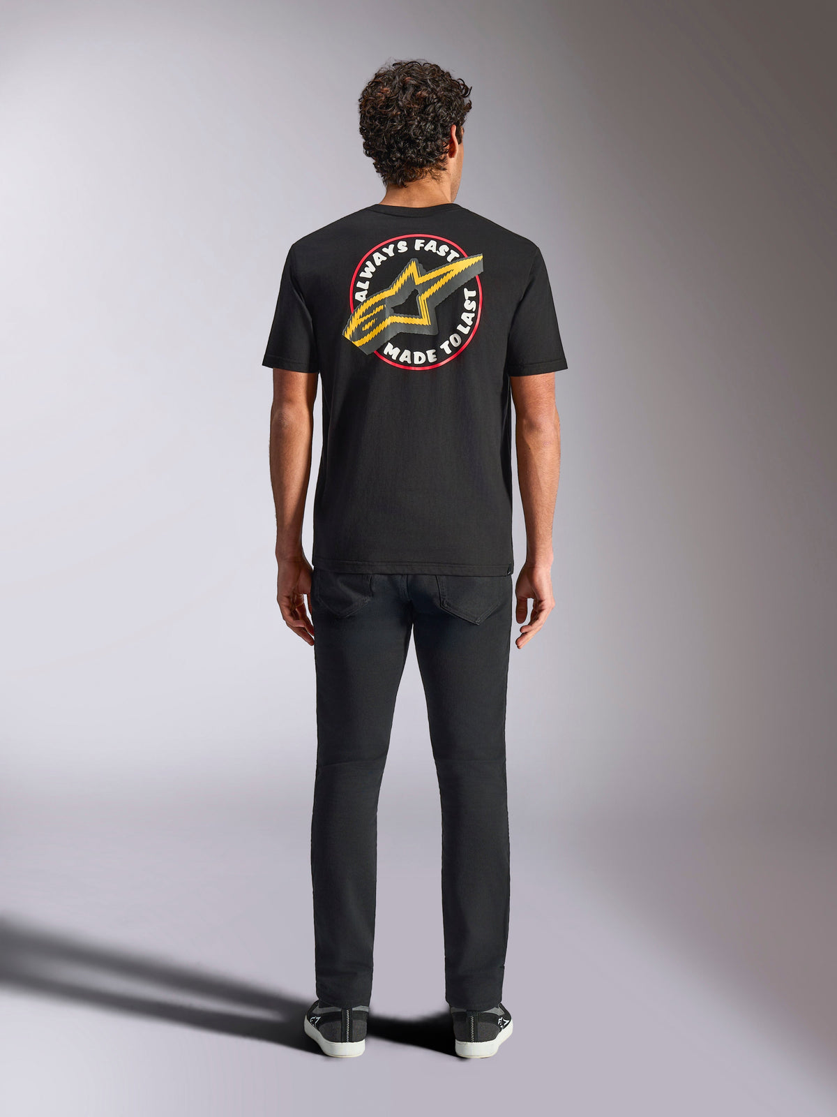 Alpinestars Aorbit CSF Tee, kortärmad t-shirt, svart, bärs av modell bakifrån med svarta byxor, med stor 'Always Fast Made to Last'-grafik med gul Astars-logotyp, avslappnad motorsport-streetwear