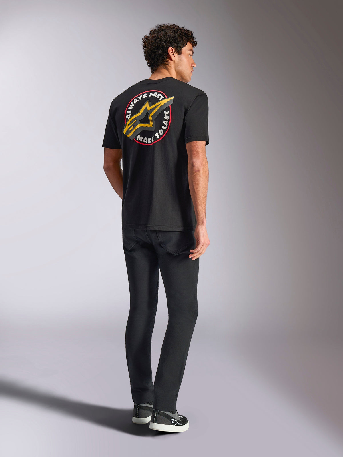 Alpinestars Aorbit CSF Tee, kortärmad t-shirt, svart, baksida med en stor gul Astars-logotyp och cirkulär grafik 'Always Fast Made to Last', buren med svarta jeans och sneakers, motorsportinspirerad streetwear-stil